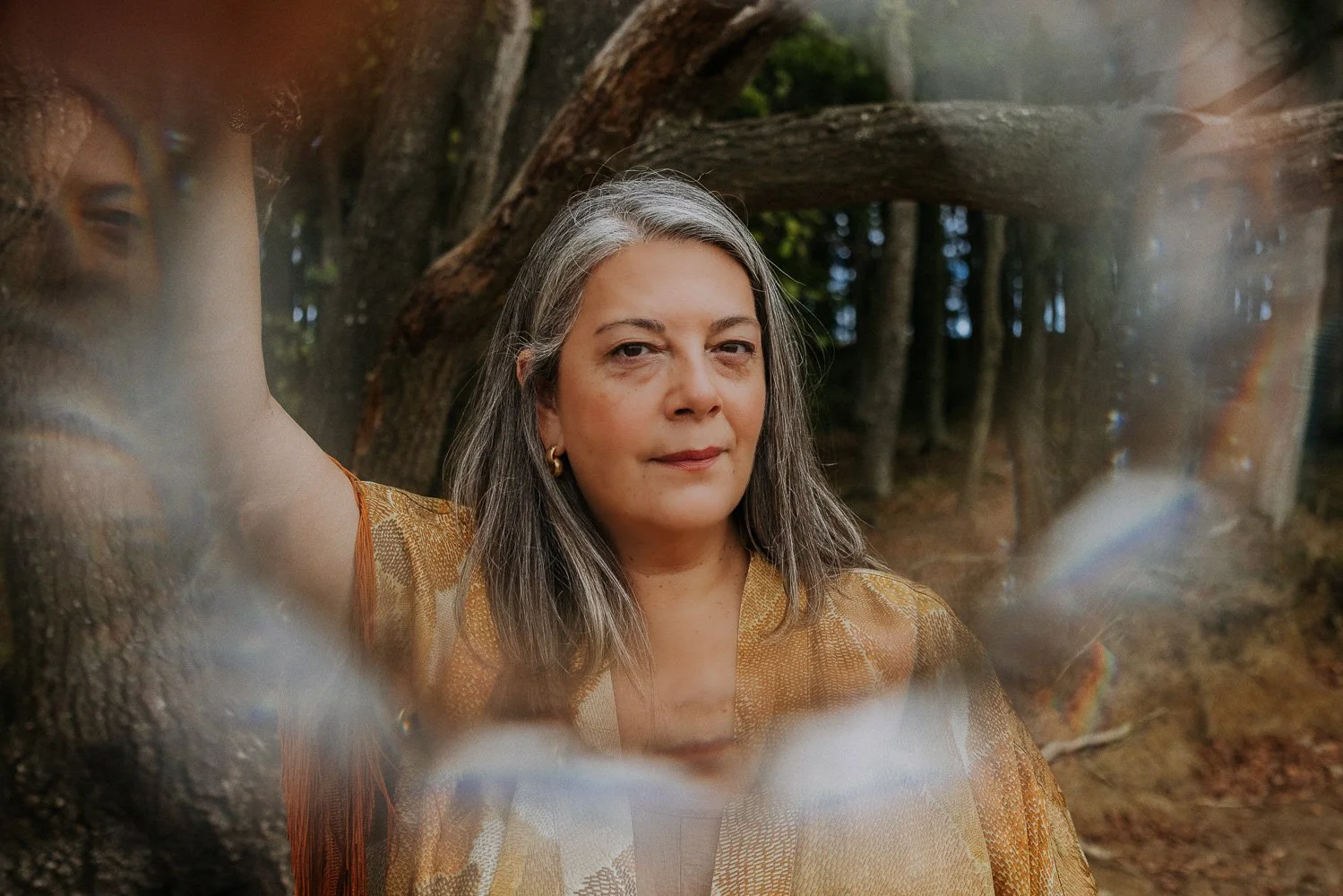 A woman with gray hair and gold earrings, wearing a patterned gold shawl, stands outdoors in a forested area, looking directly at the camera with a calm expression. She is framing herself with her arm and the shot includes natural light and trees in the background.