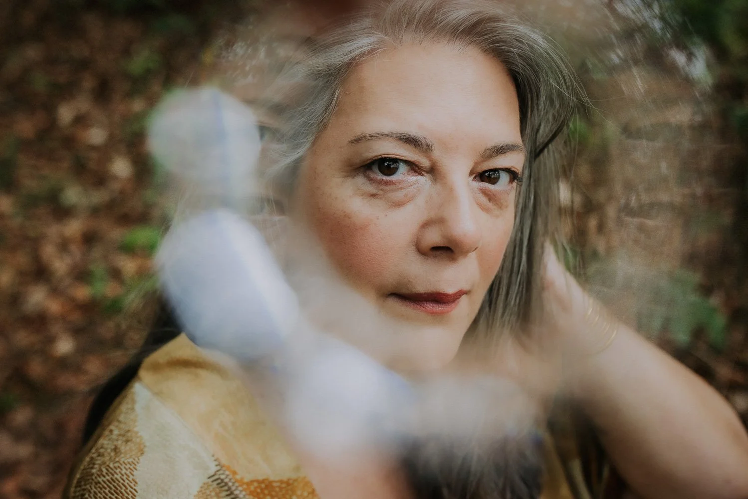 A woman with gray hair and freckles resting her head on her arm outdoors with a blurred background of fallen leaves and green foliage.