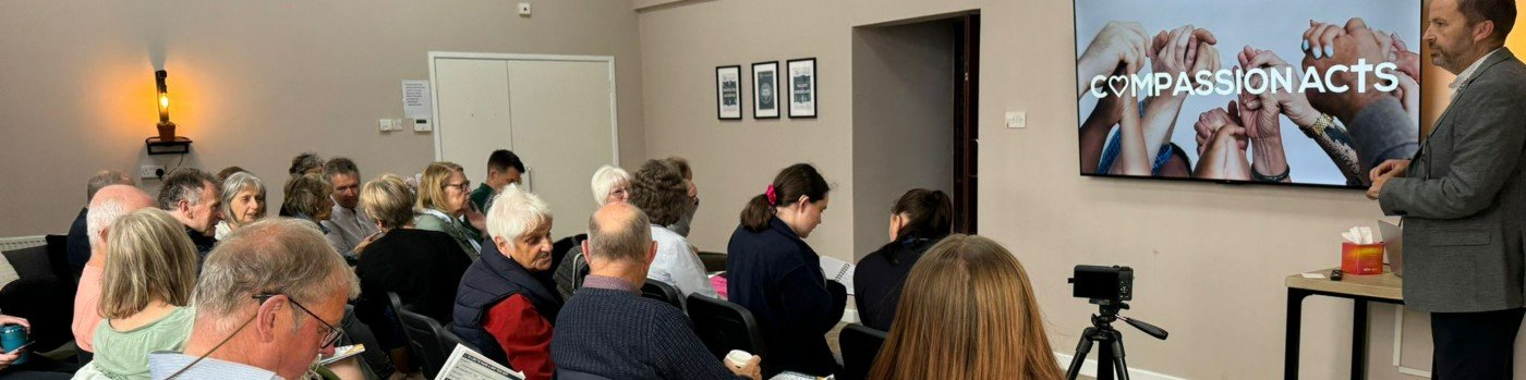 A group of people attending a presentation or seminar in a conference room. A man in a gray suit is standing on the right side near a screen that displays a hands clasped together with the text 'COMPASSION ACTS'.