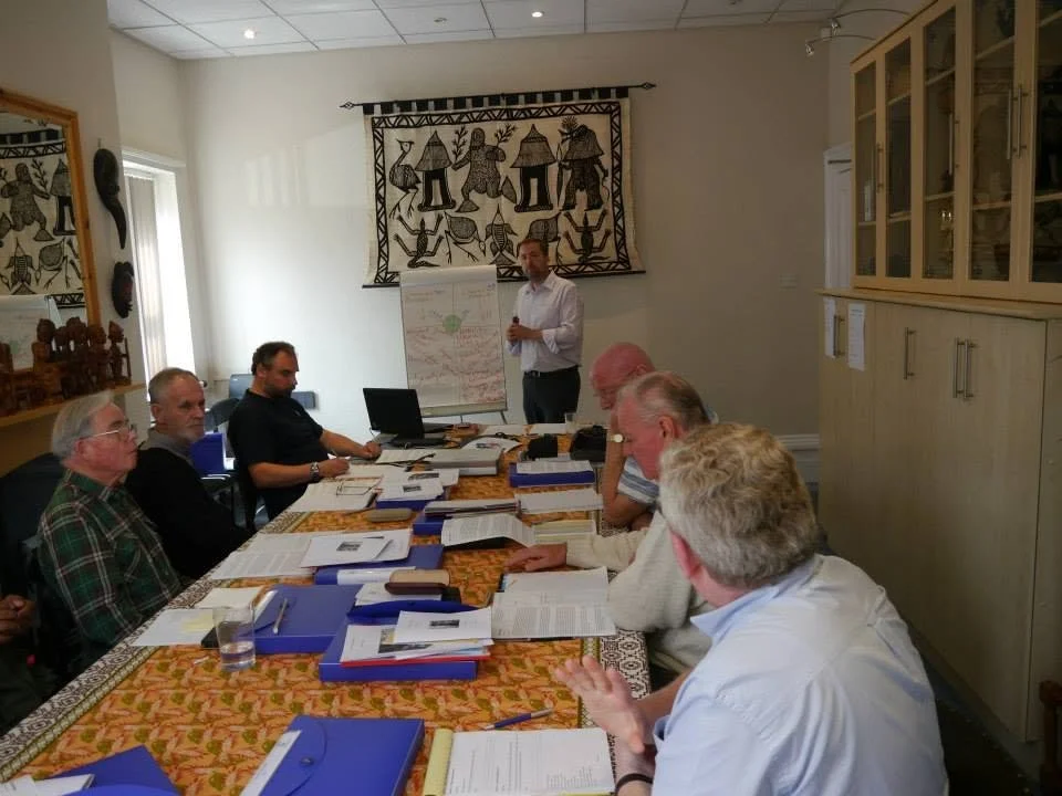 A man standing in front of a whiteboard giving a presentation to a group of people seated at a table in a conference room.