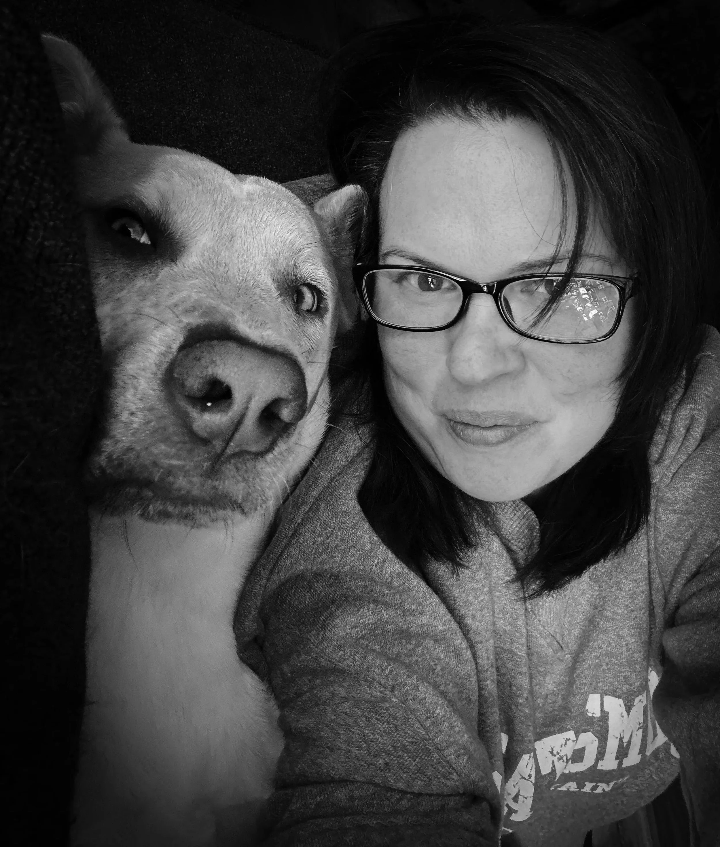 Black-and-white photo of a person with glasses and dark hair lying close to a light-colored dog with a short fur coat. The person's face is near the dog's face with their eyes visible, and the dog appears relaxed.