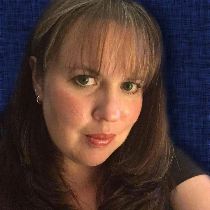 A woman with shoulder-length brown hair and green eyes, wearing earrings and a black top, sitting against a dark blue background.