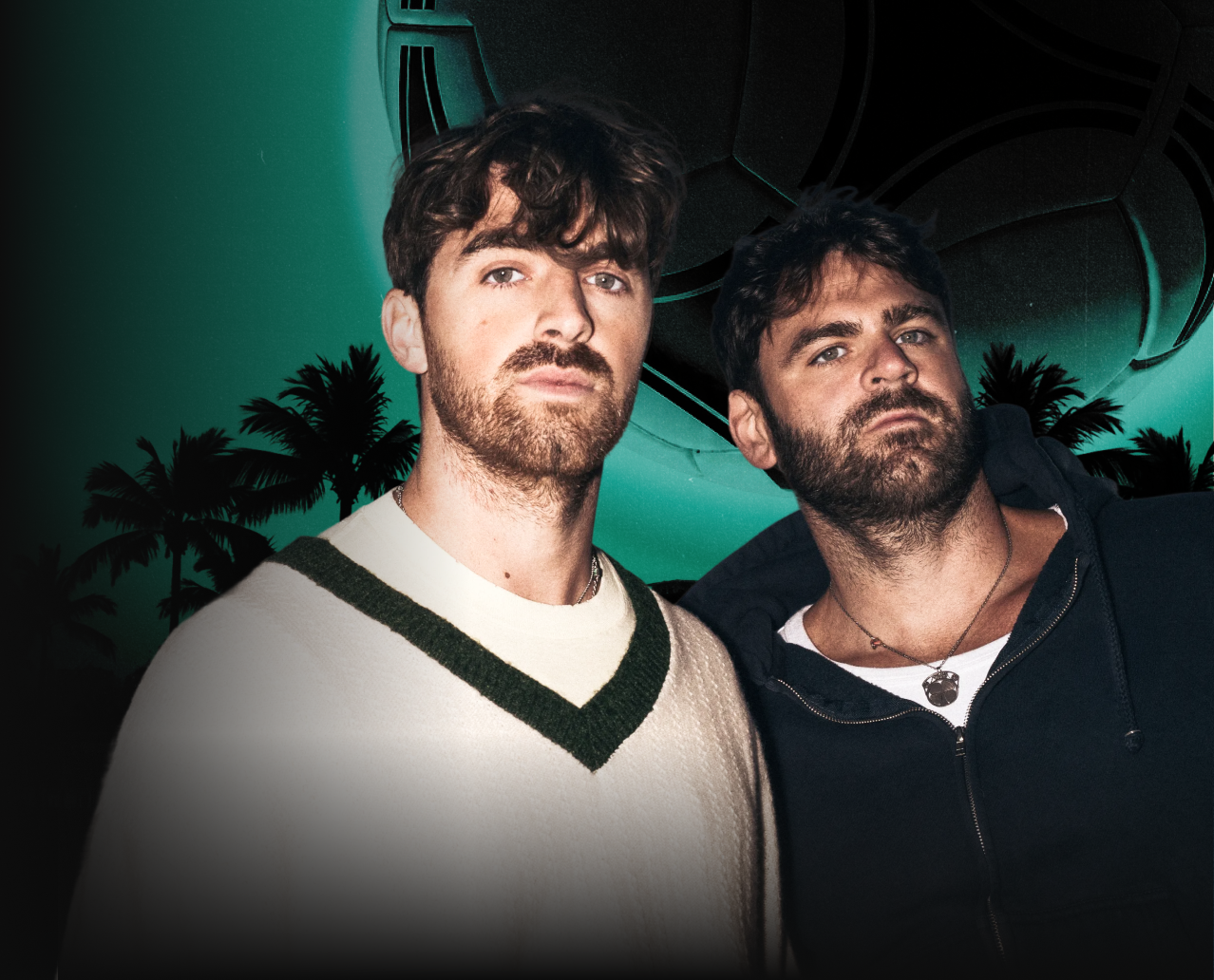 Two men with beards and dark hair standing outdoors during dusk, with palm trees and a large structure in the background.