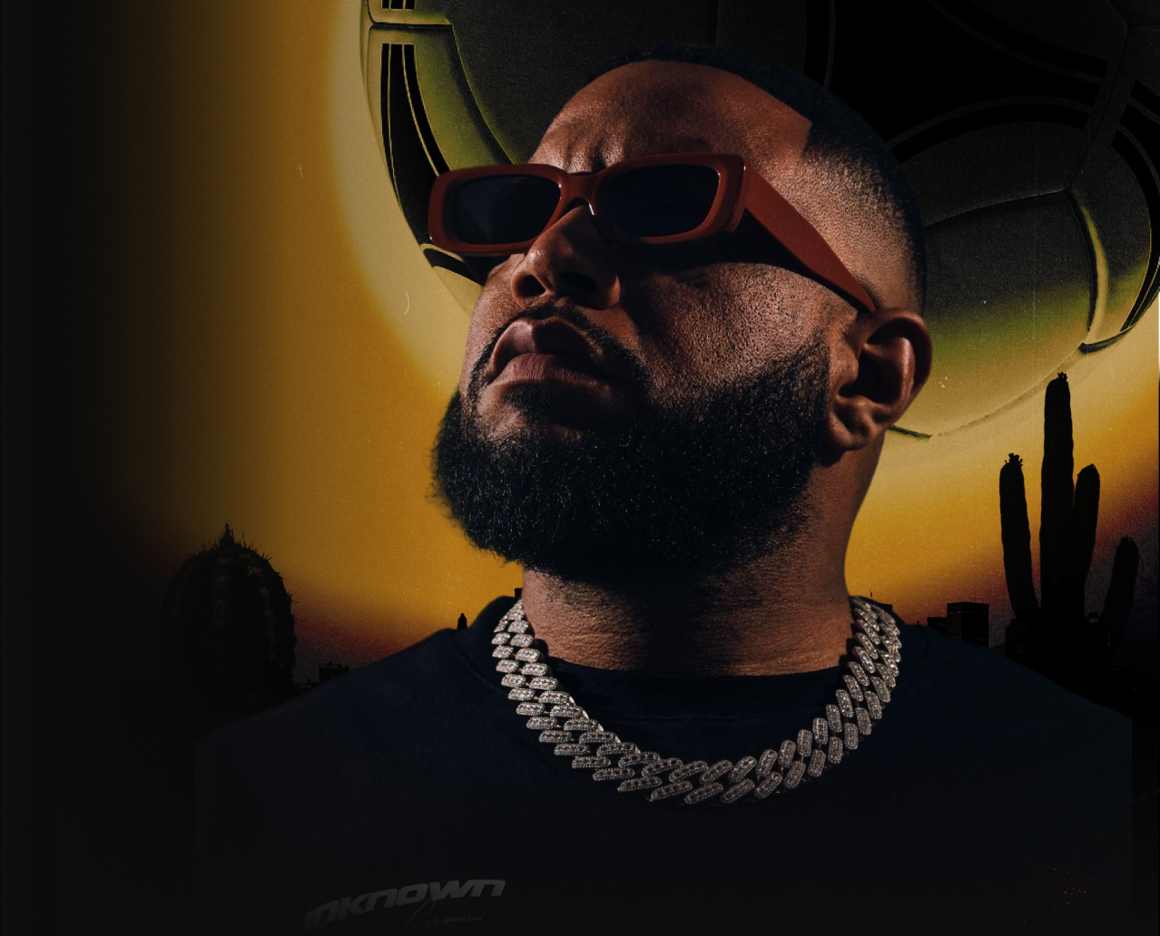 A man with a beard wearing large sunglasses, a thick chain necklace, and a black shirt with a cityscape and desert plants in the background at sunset.