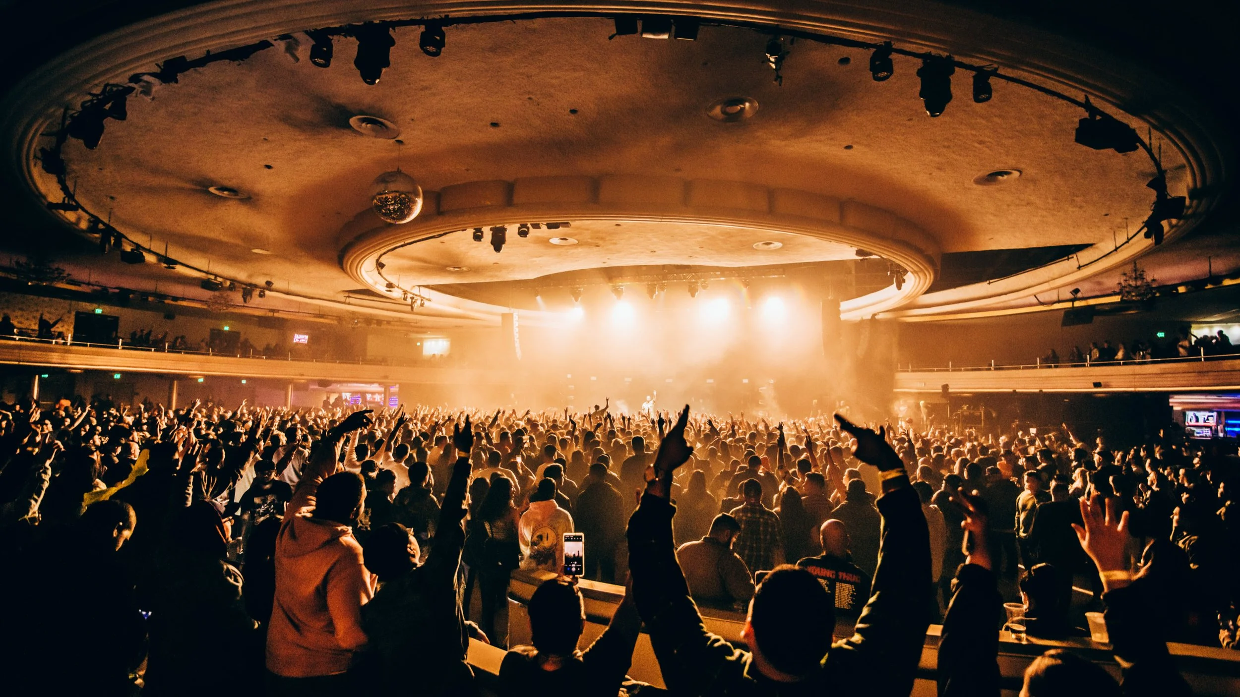 Crowd at a concert with bright stage lights, a disco ball, and audience raising their hands.