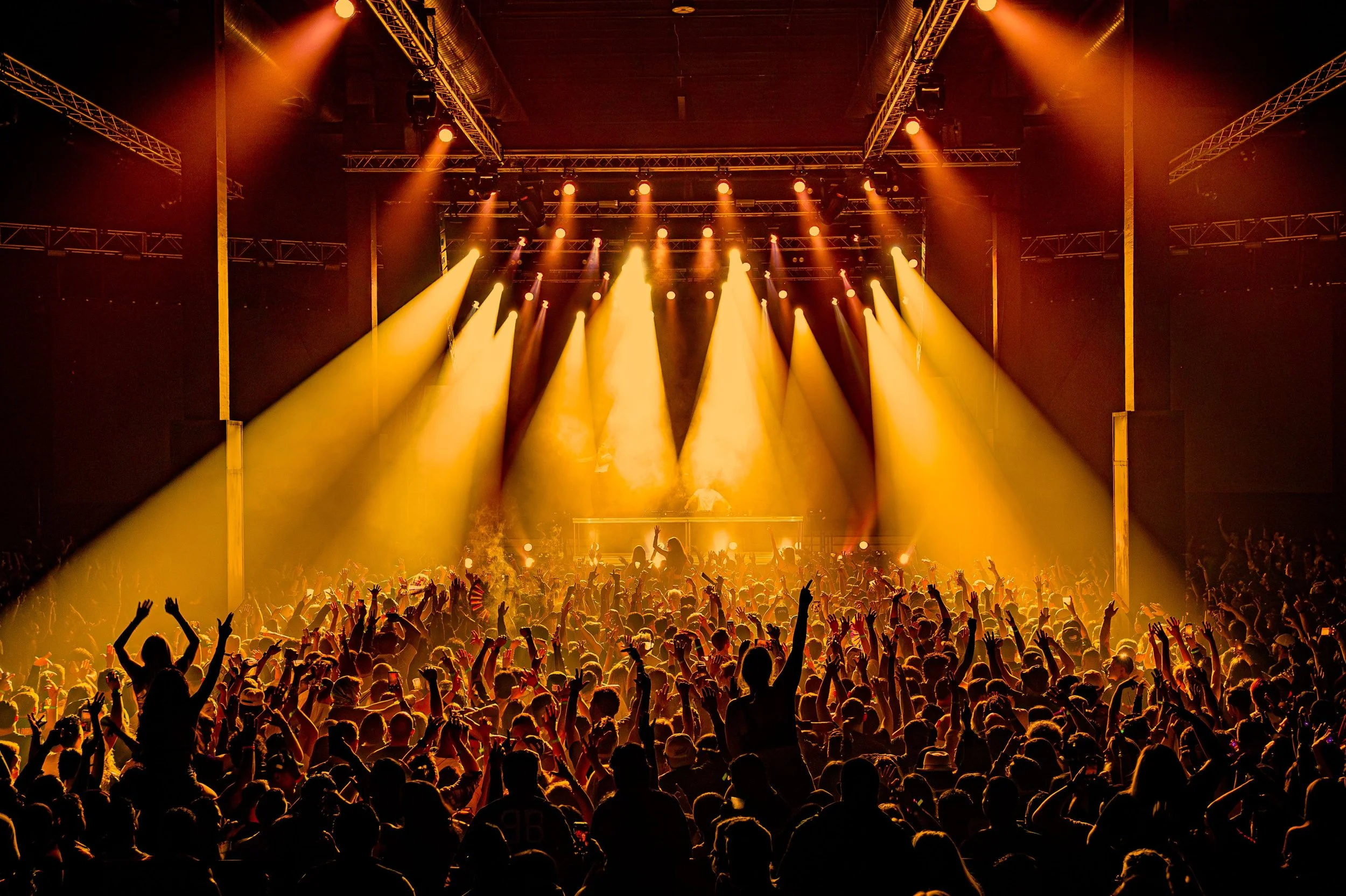 A concert with a large crowd dancing under bright yellow and orange stage lights and rigging.