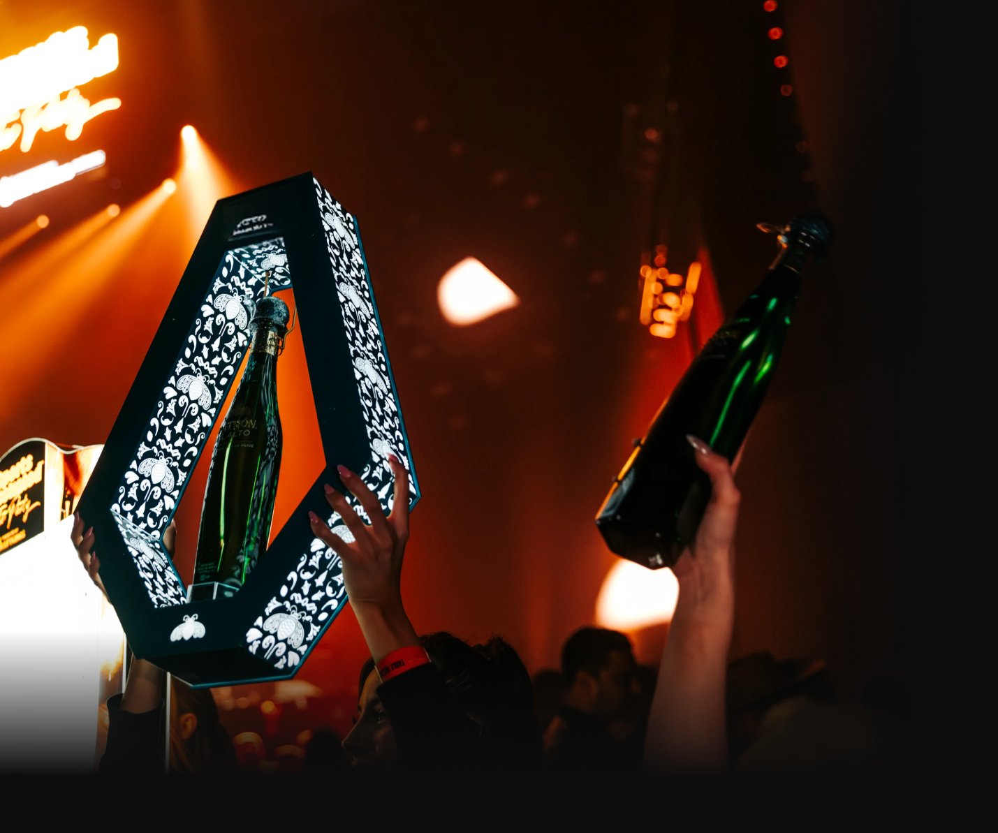 People holding up bottles inside decorative frames at an event with dark background and orange lighting.