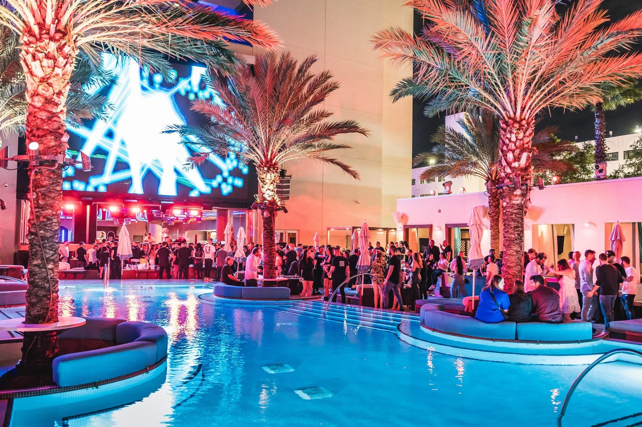 Night scene at a poolside party with many people, palm trees lit with pink and blue lights, and a large digital screen in the background showing abstract blue and white graphics.
