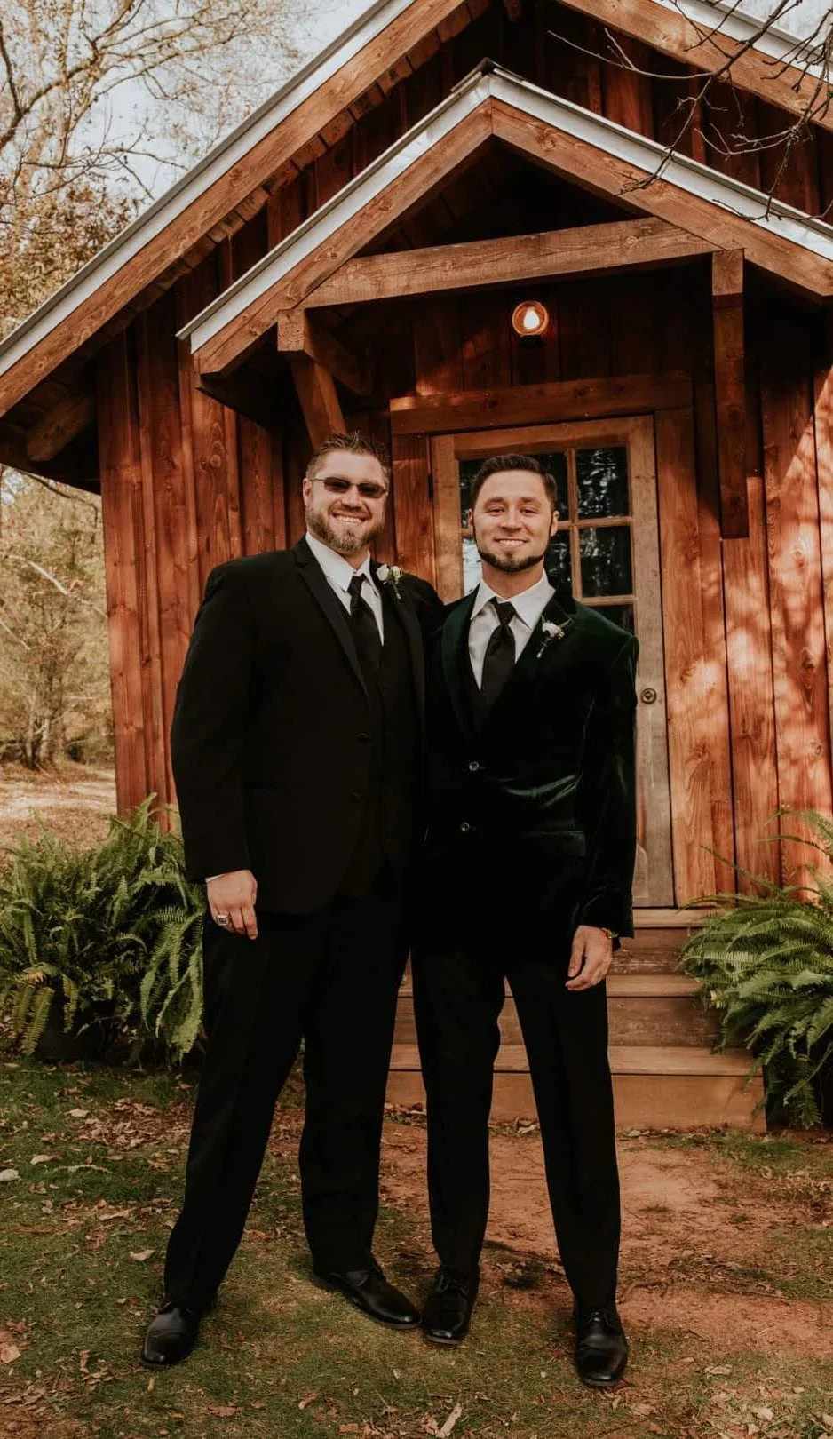 Two men in black tuxedos standing outside a small wooden house with a porch and surrounded by greenery.