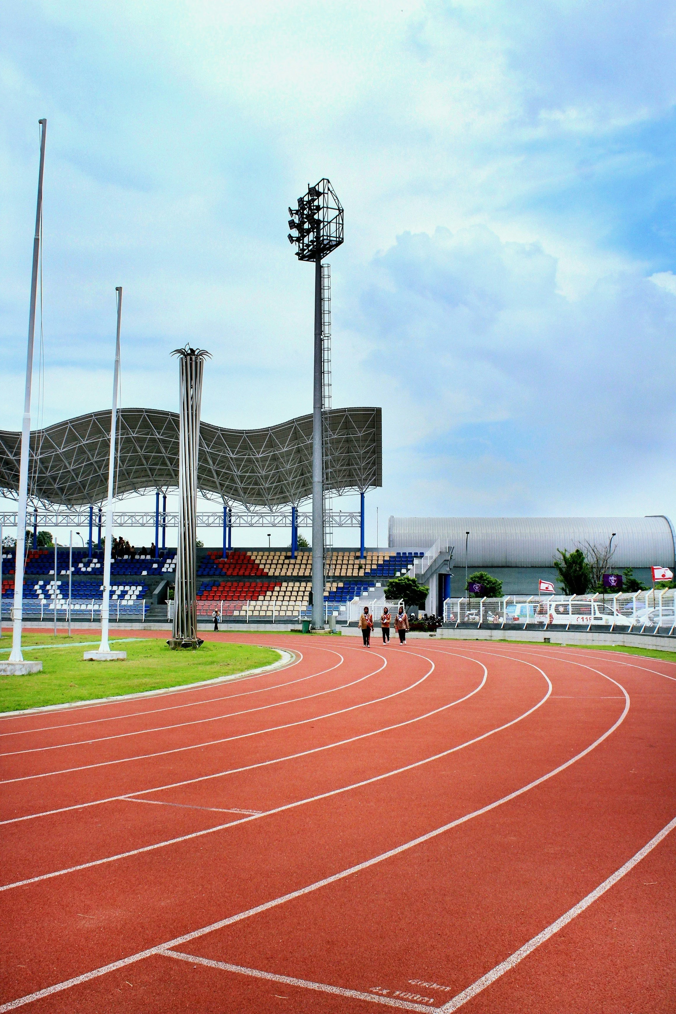 An outdoor sports stadium with a running track and seating area, a scoreboard tower, and a canopy covering some seats, under a cloudy sky.