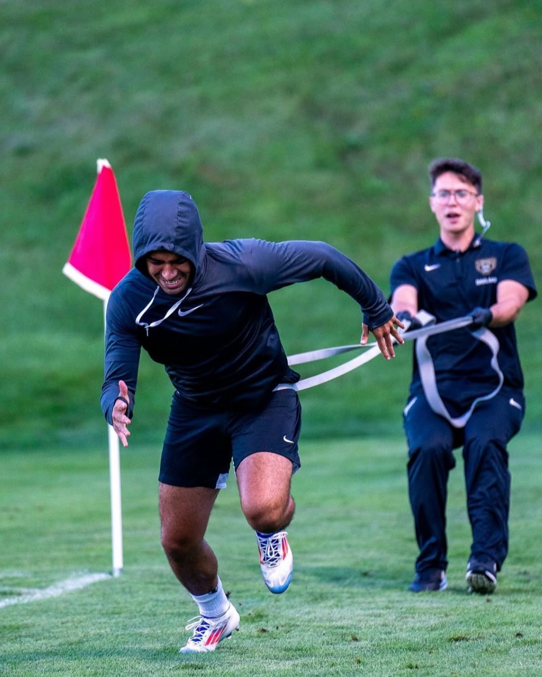 A male athlete in a hoodie and shorts running on a grassy field, pulling a resistance band with a person in the background holding the other end, near a red flag on the field.