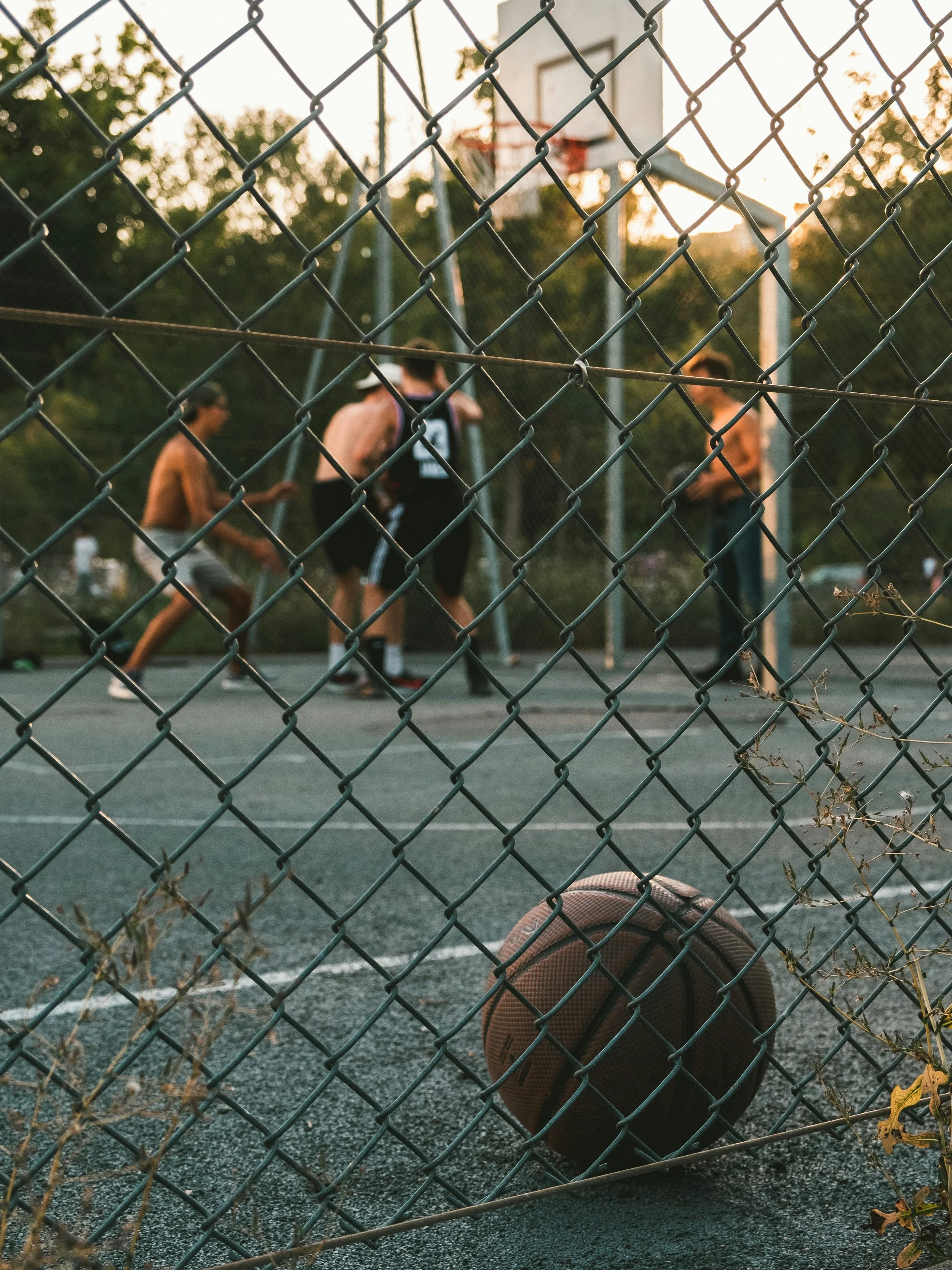 A basketball on a sports court, seen through a chain-link fence, with several people playing basketball in the background during sunset.