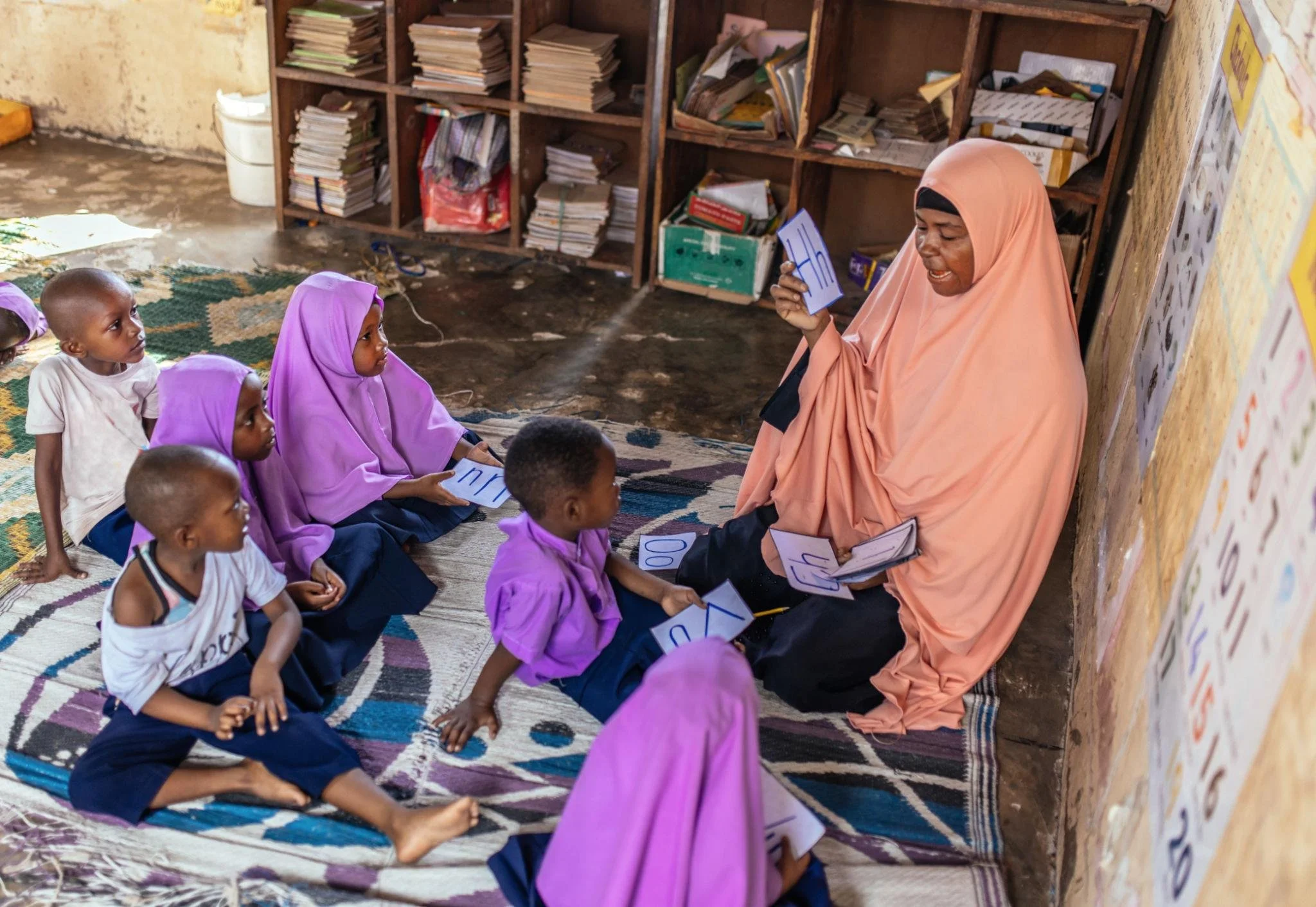A woman wearing a peach hijab sitting on the floor in front of a blackboard, teaching a group of young children who are sitting on a multicolored rug. The children hold cards with letters, engaging in a learning activity inside a classroom with wooden shelves filled with books and papers in the background.
