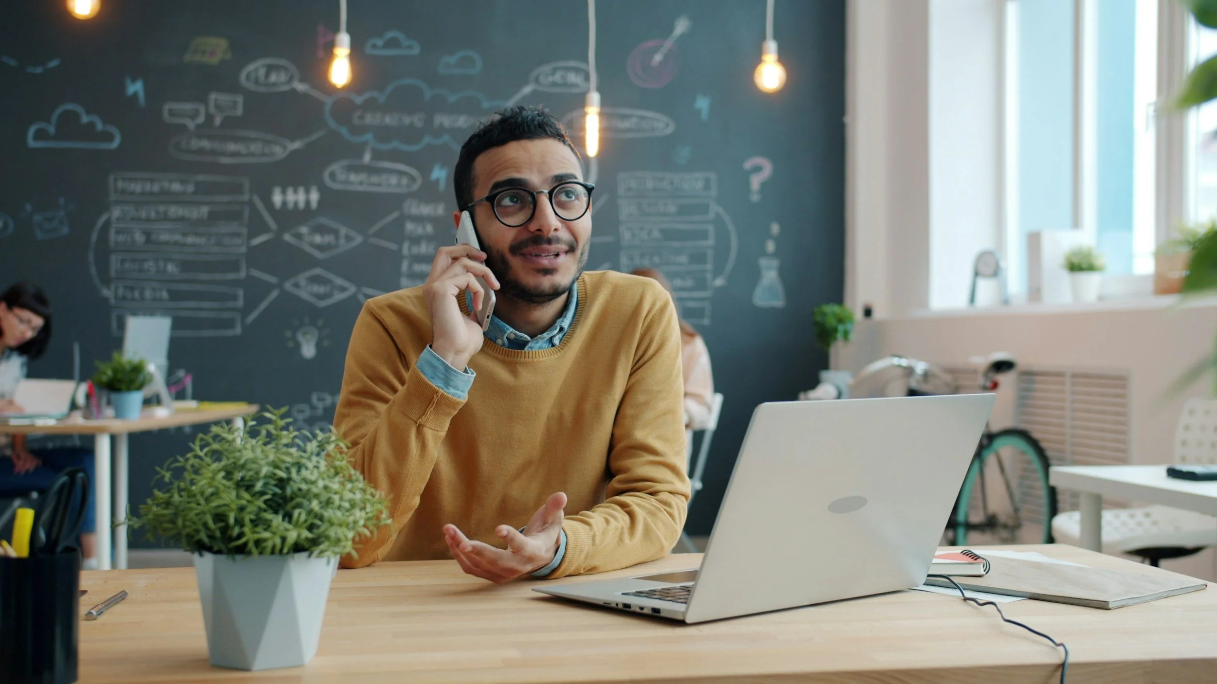 A man with glasses, wearing a mustard-colored sweater, talking on a mobile phone while sitting at a desk with a laptop and a potted green plant. In the background, a chalkboard wall has various drawings and notes, with bright natural light flowing in from windows.