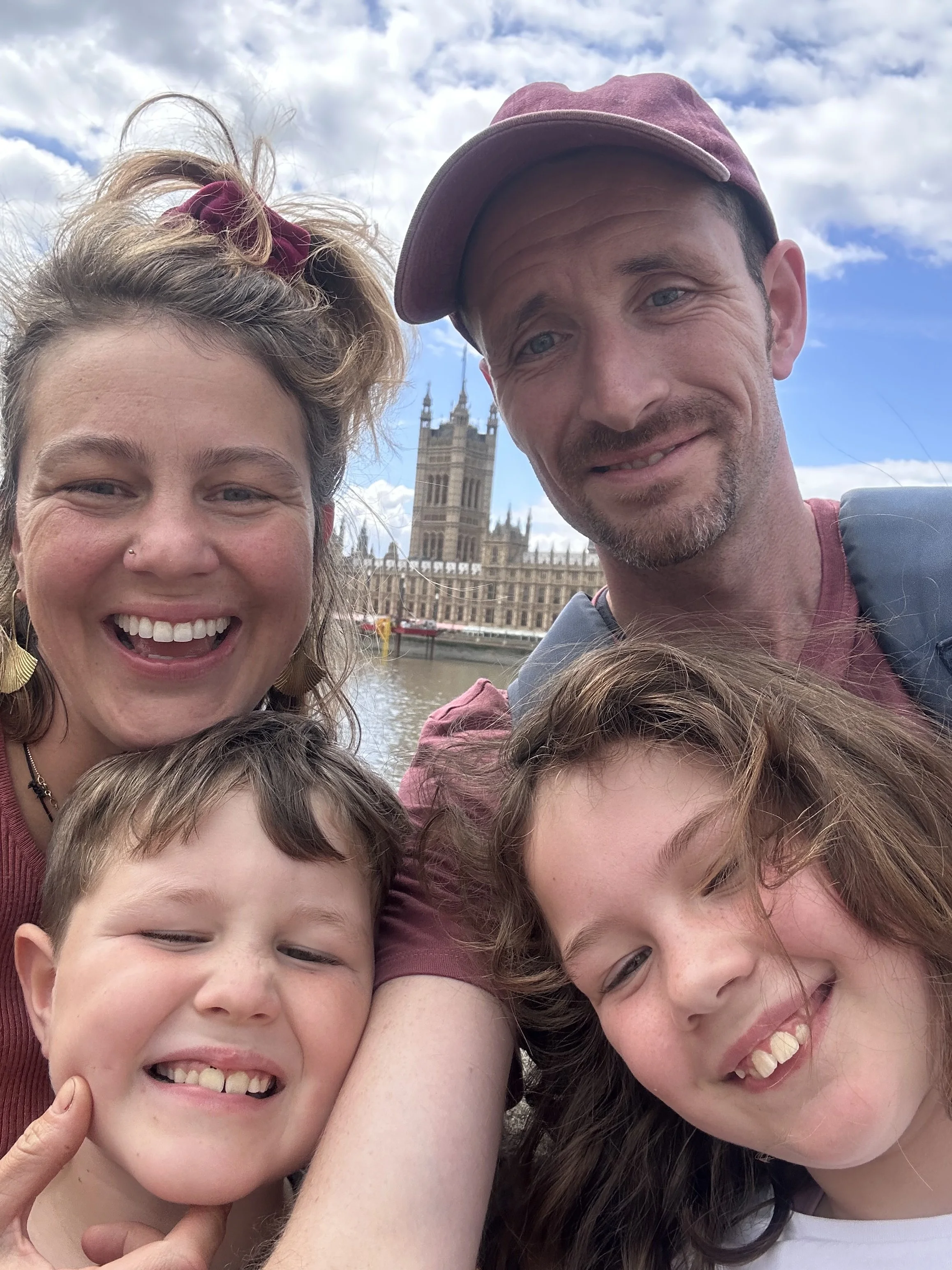 A group selfie of smiling people, including children and adults, with the Big Ben clock tower and a cloudy sky in the background.