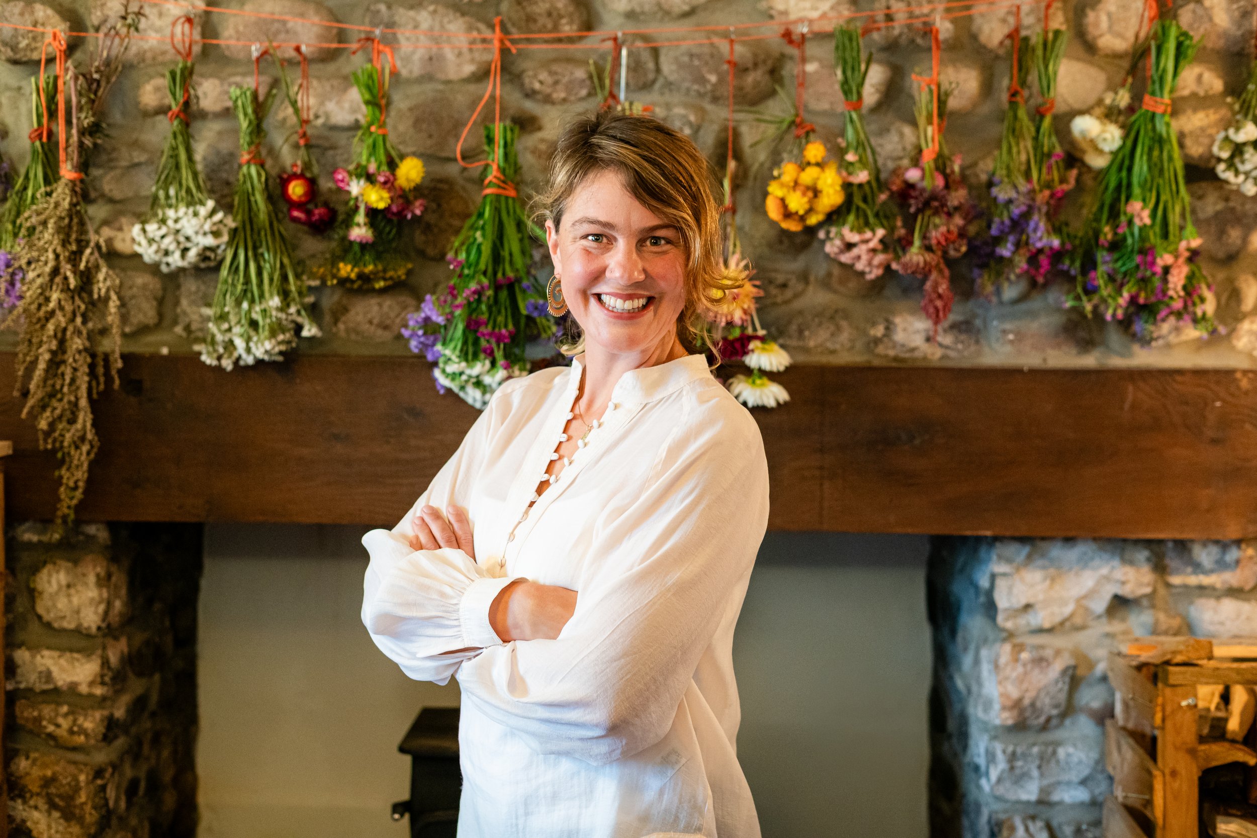 A woman with short brown hair smiling and crossing her arms, standing in front of a stone fireplace with hanging bunches of colorful flowers tied with orange ribbons.