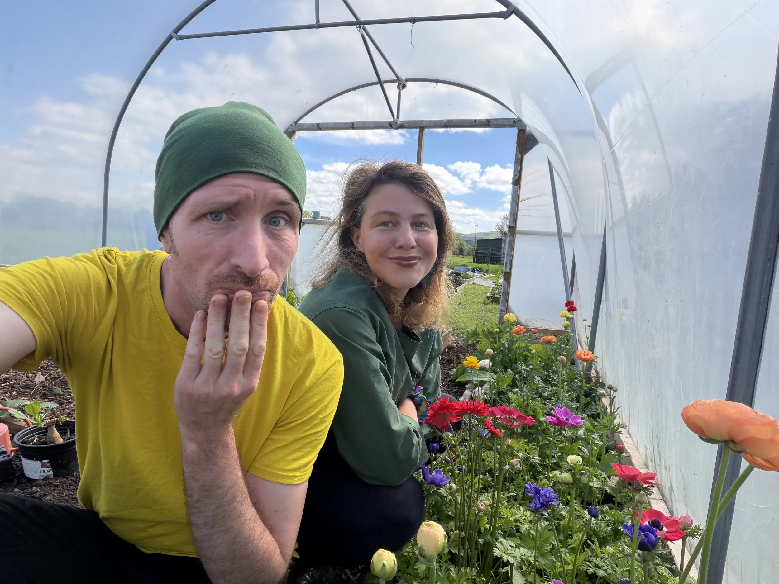 Two people, a man wearing a green head covering and a yellow shirt, and a woman with long hair in a green jacket, inside a greenhouse with a variety of colorful flowers, including red, purple, and yellow blossoms, on a sunny day with blue skies and clouds.