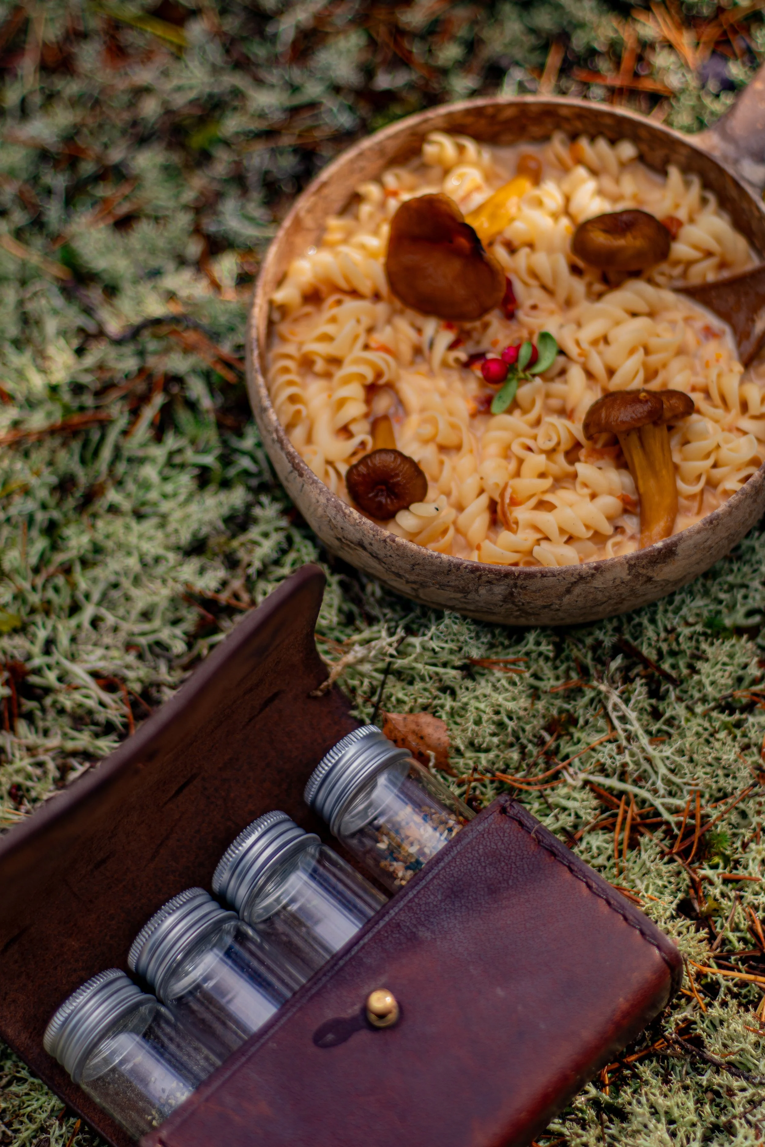 A wooden bowl filled with pasta and mushrooms, placed on mossy ground outdoors. To the lower left, there's a leather case holding several small glass jars with metal lids. The scene has a natural, rustic feel.