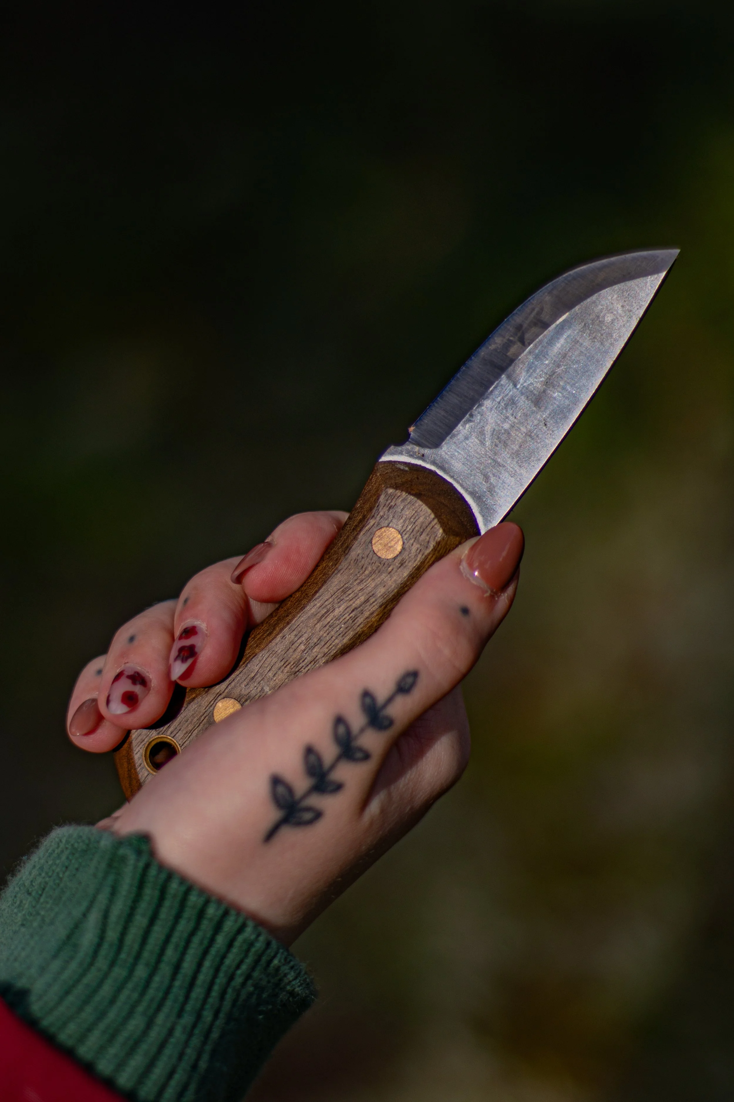 Close-up of a hand with painted nails holding a small hunting knife with a wooden handle against a blurred outdoor background.