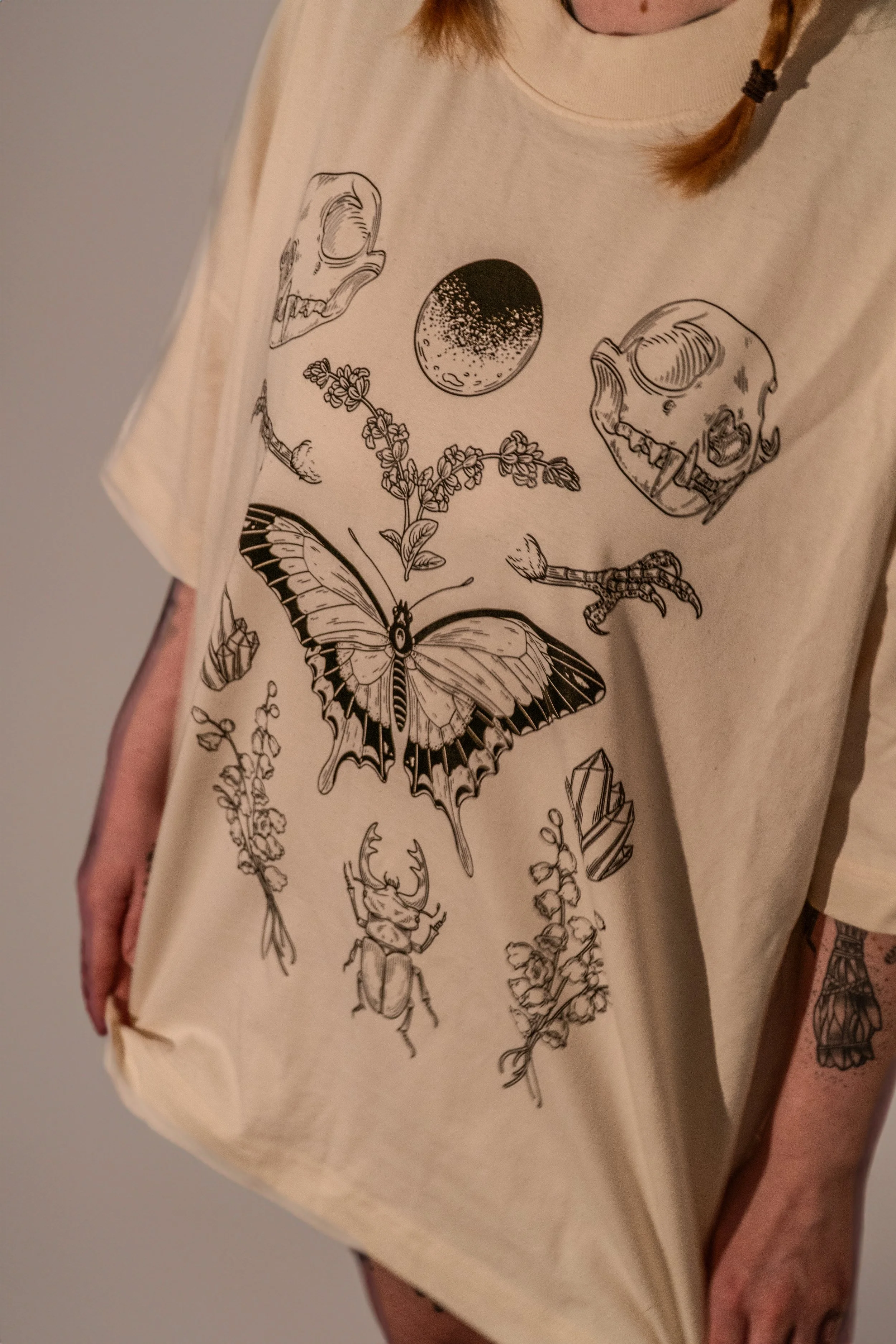 Person wearing a beige shirt with black line drawings of skulls, butterflies, flowers, a moon, and an animal skull. Part of person's arm and tattoo are visible.