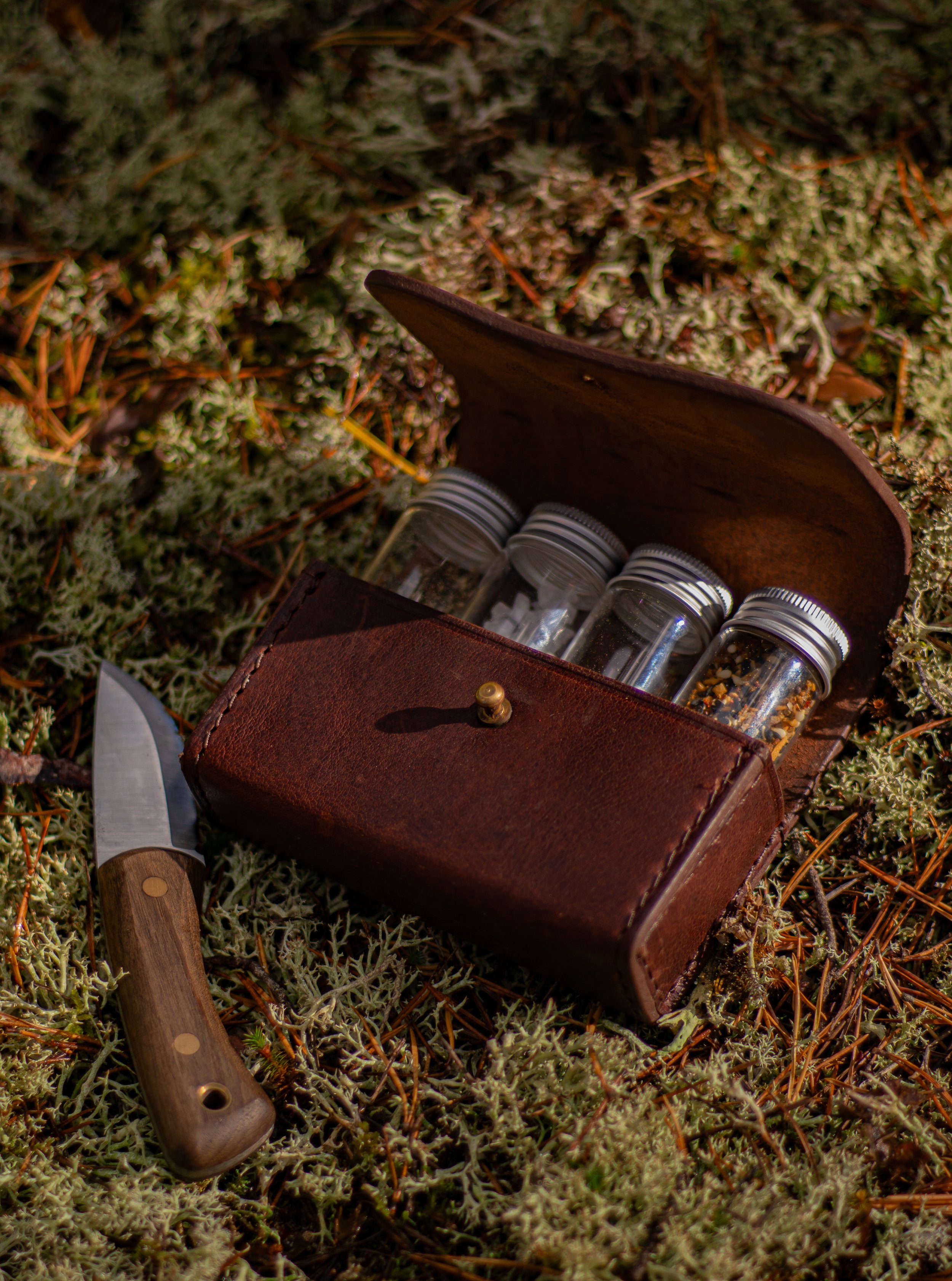 A leather case containing small glass jars with black and white caps, situated on mossy ground, with a small wooden-handled tool nearby.