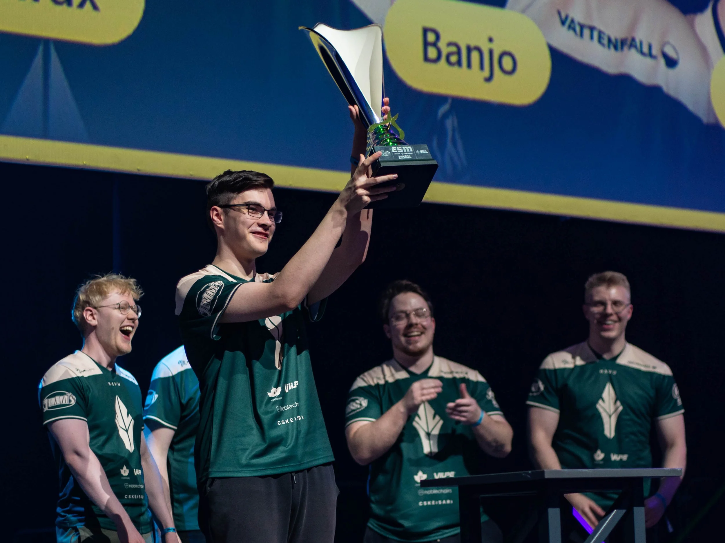 A man in a striped green and white sports jersey holding a trophy on a stage, surrounded by smiling teammates.