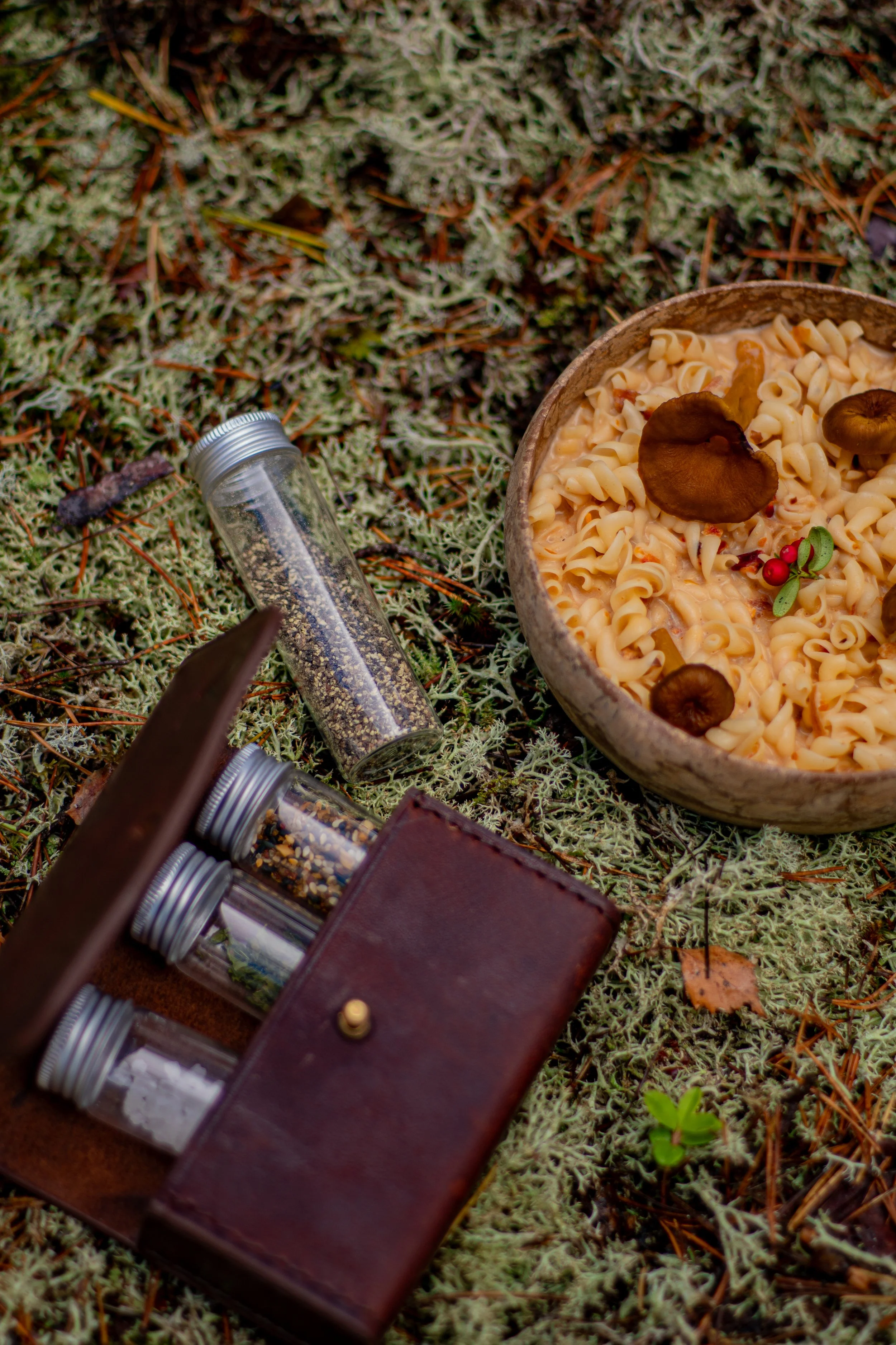 A bowl of pasta with mushrooms and herbs, surrounded by small glass jars of spices, in an outdoor natural setting.