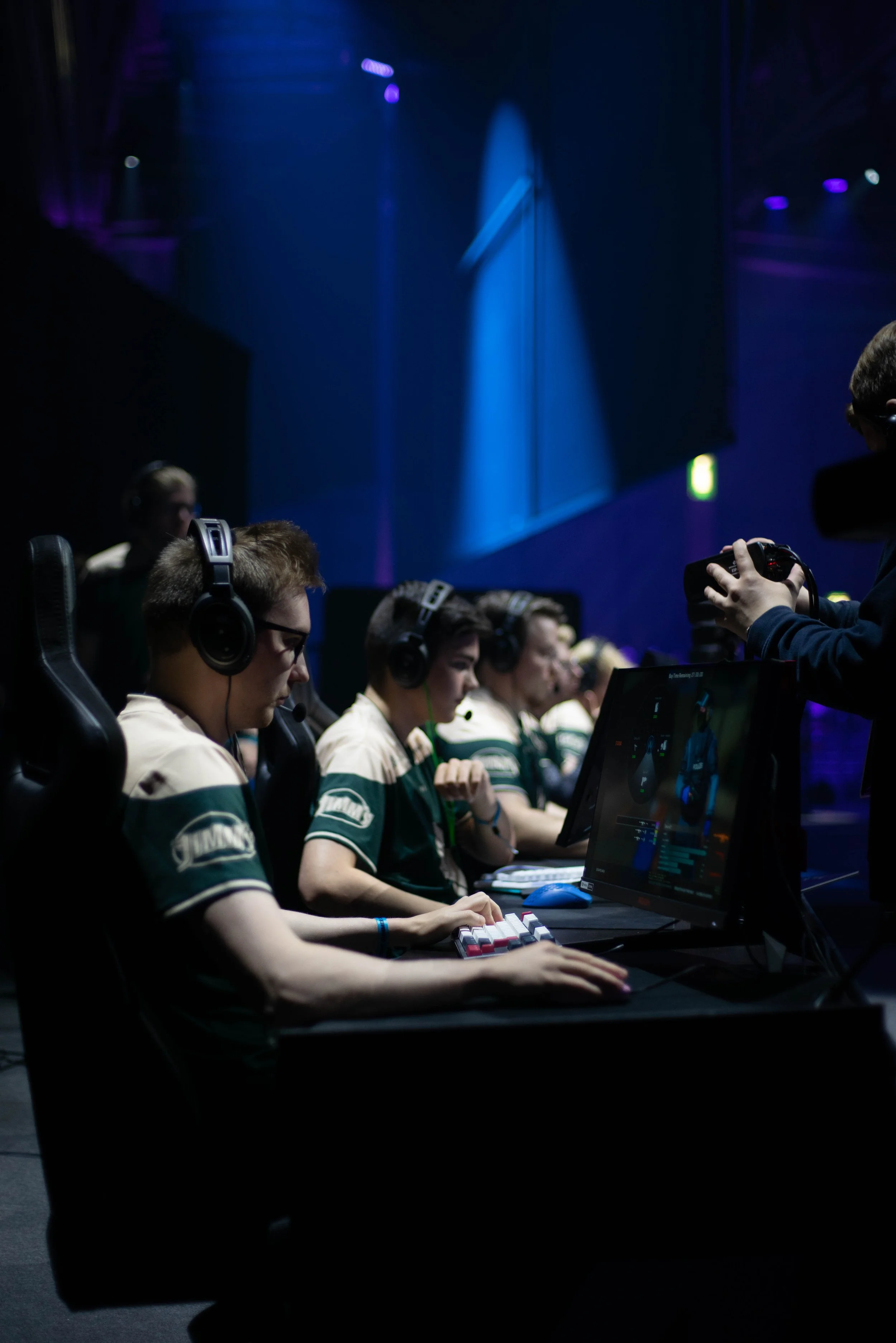 A group of young people participating in an esports or gaming tournament, wearing headphones and focusing on their computer screens, with a dark stage and blue lighting in the background.