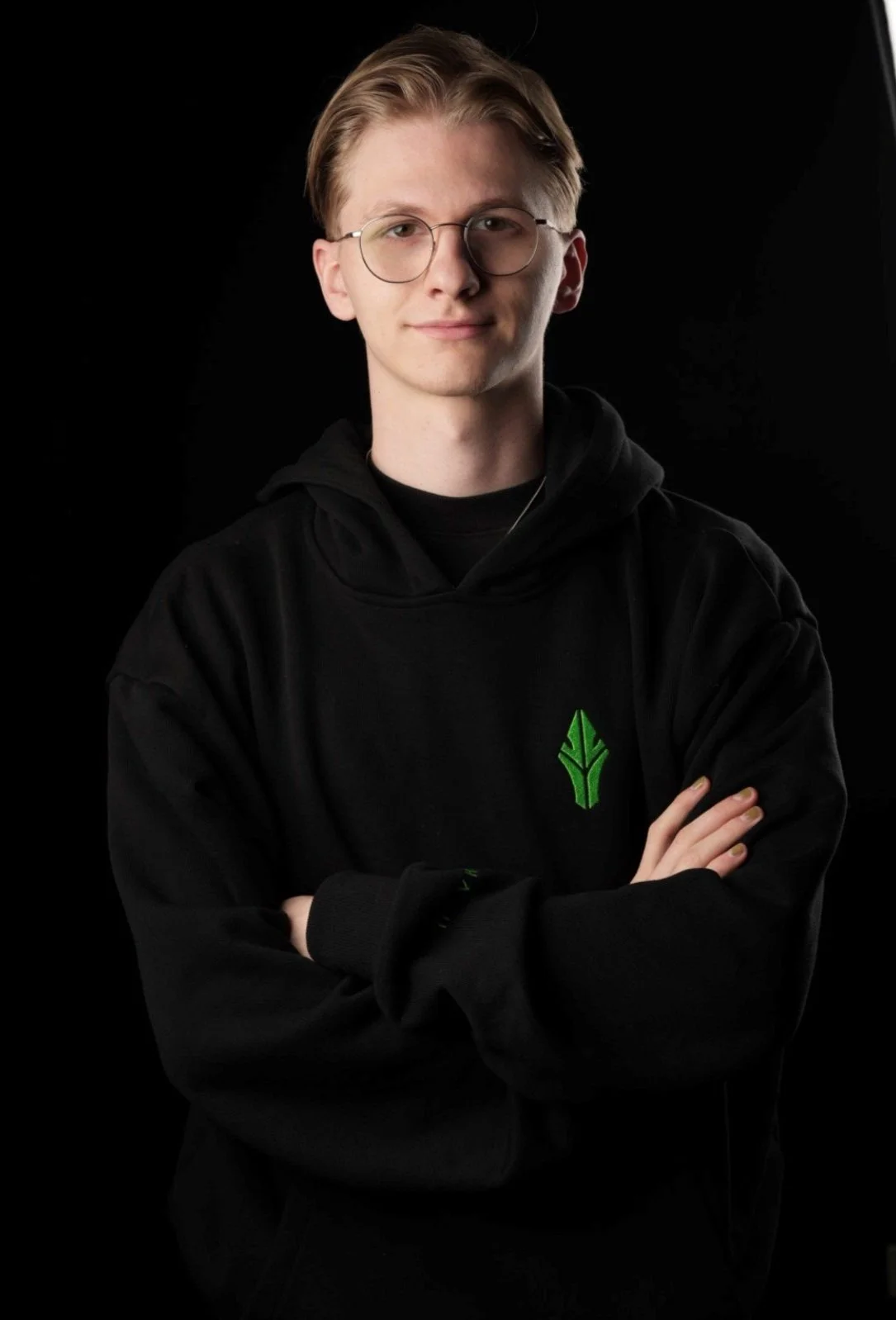 A young man with blond hair, glasses, and a black hoodie with a green logo, standing with arms crossed against a black background.