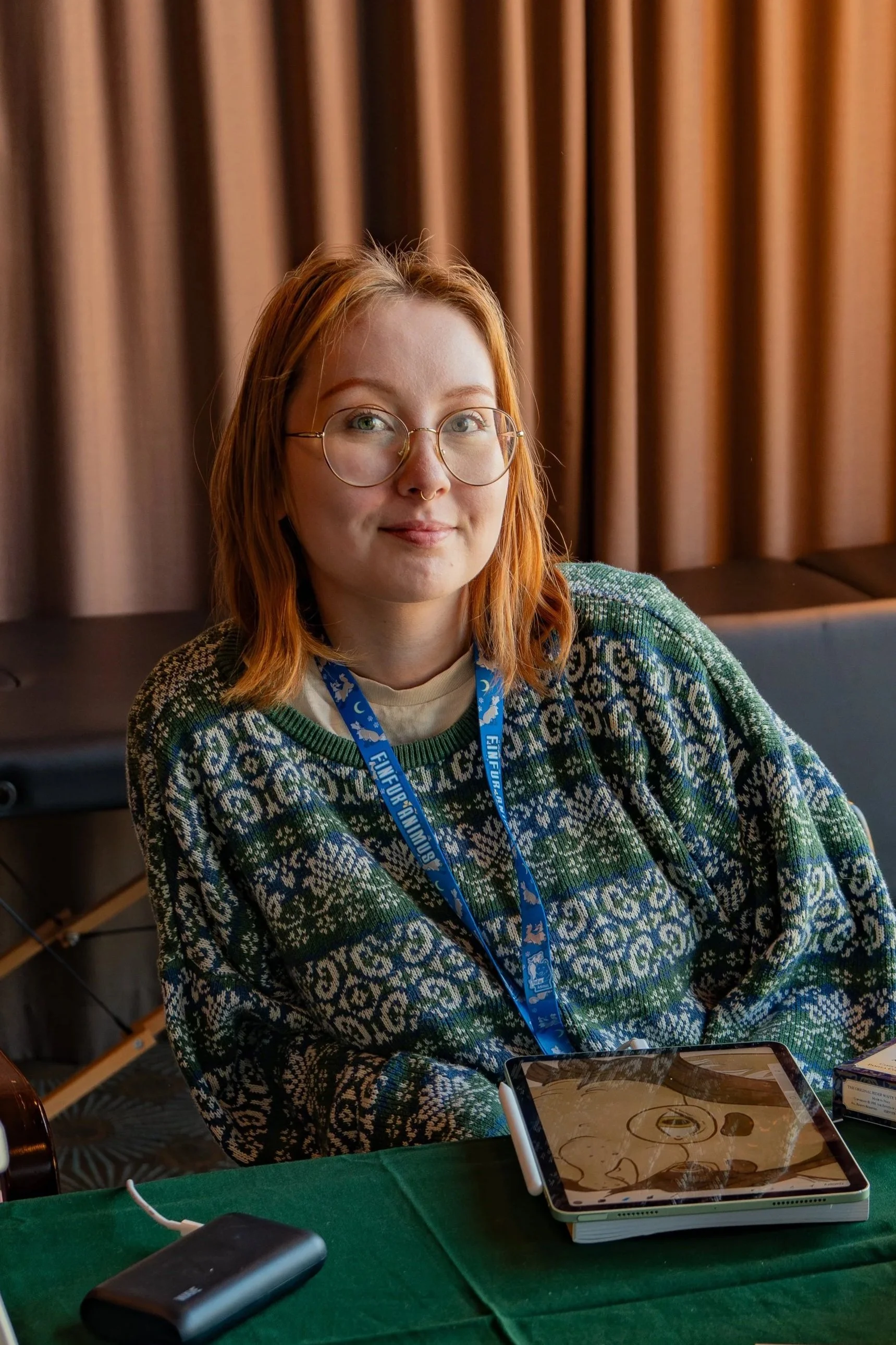 A young woman with red hair and glasses sitting at a table with a green tablecloth, a tablet, and other electronic devices, in front of brown curtains, wearing a Christmas sweater and a lanyard.