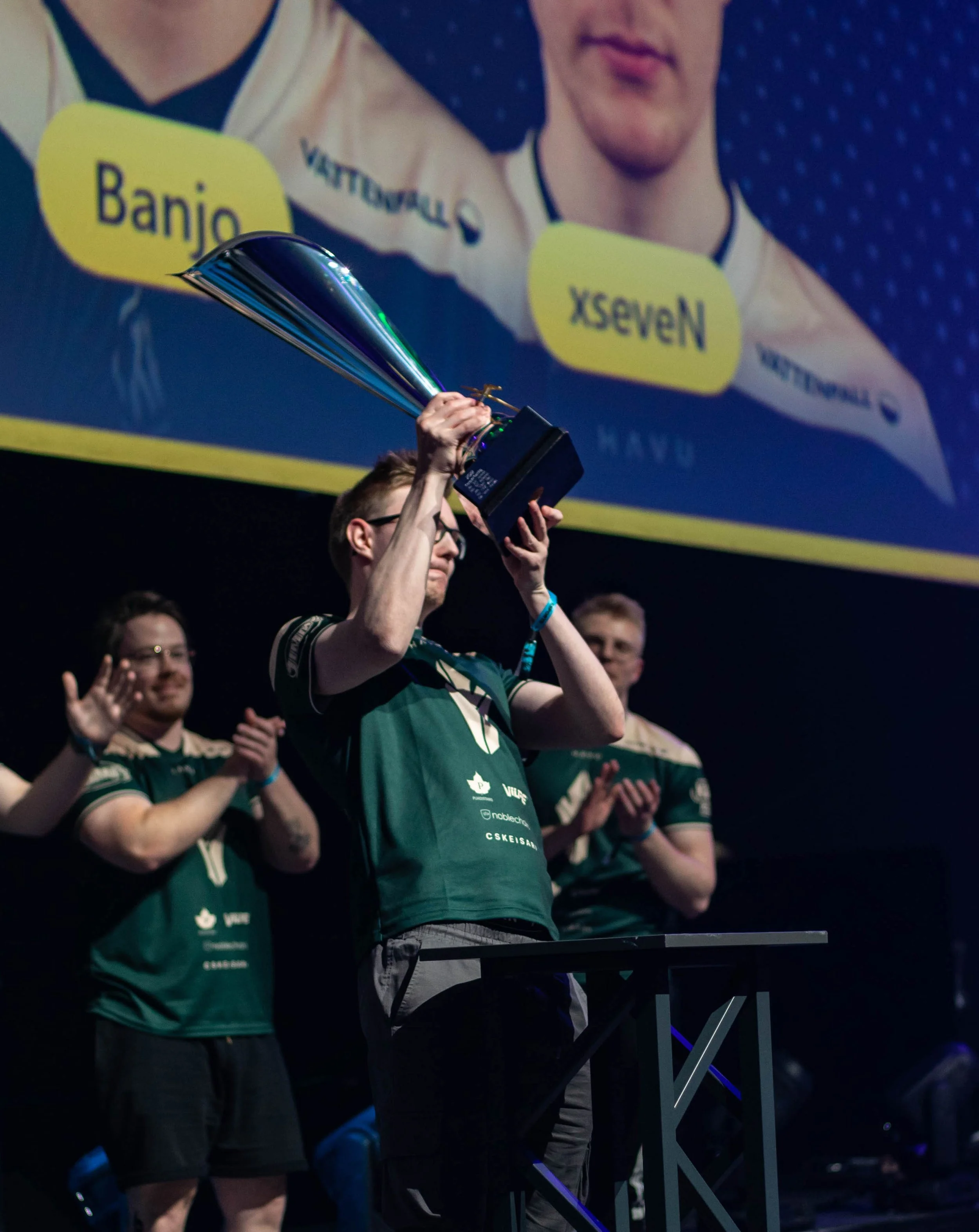 A person holding a large trophy on stage with a screen in the background displaying a close-up of their face and the text 'Banjo' and 'XSeven.' The person and others are wearing matching dark green jerseys with logos. The stage is dark with blue ligh