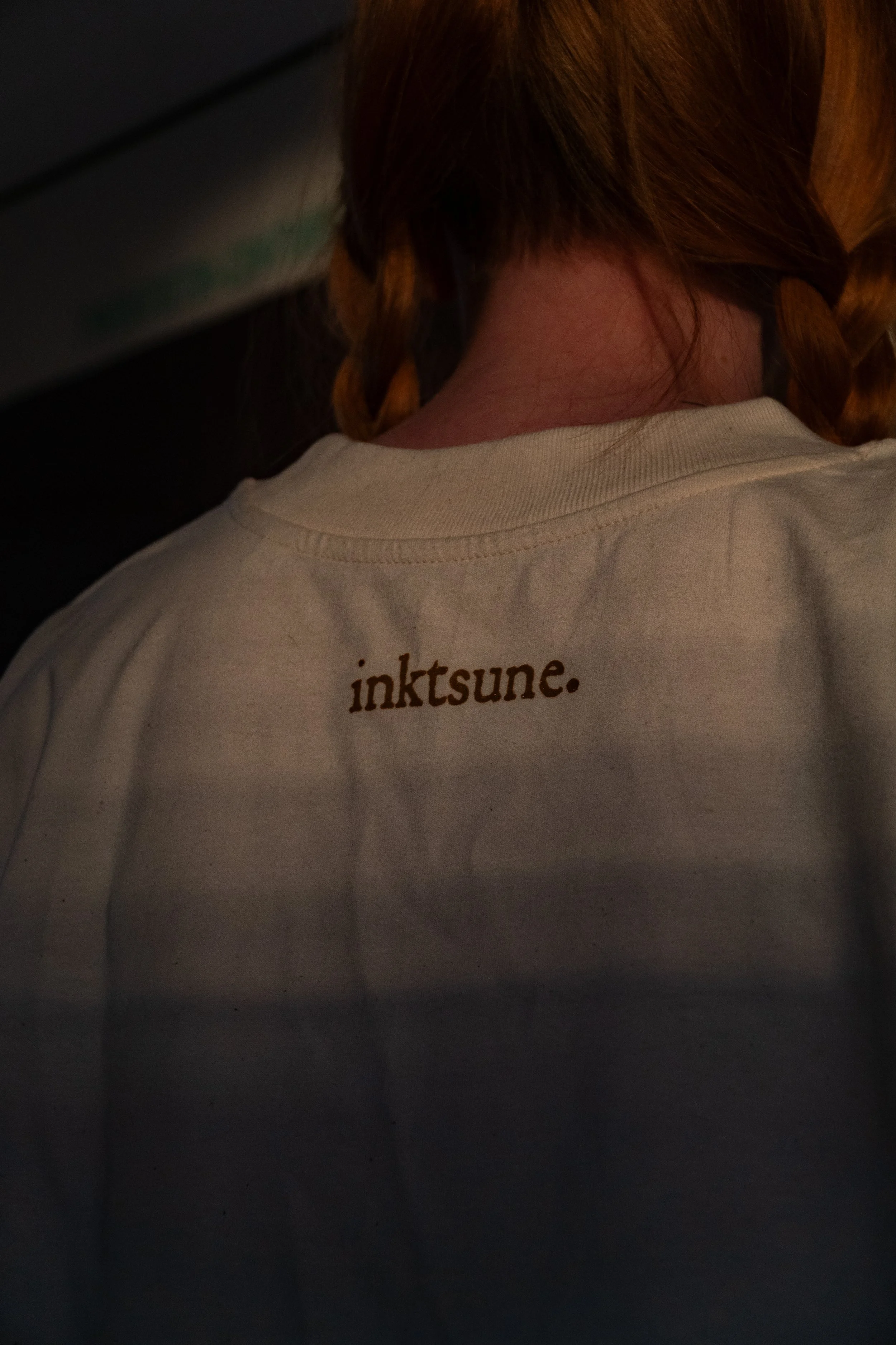 Close-up of a person with red hair and braids wearing a white shirt with the word 'inktsune.' printed on the back, seen from behind, in a dimly lit environment.