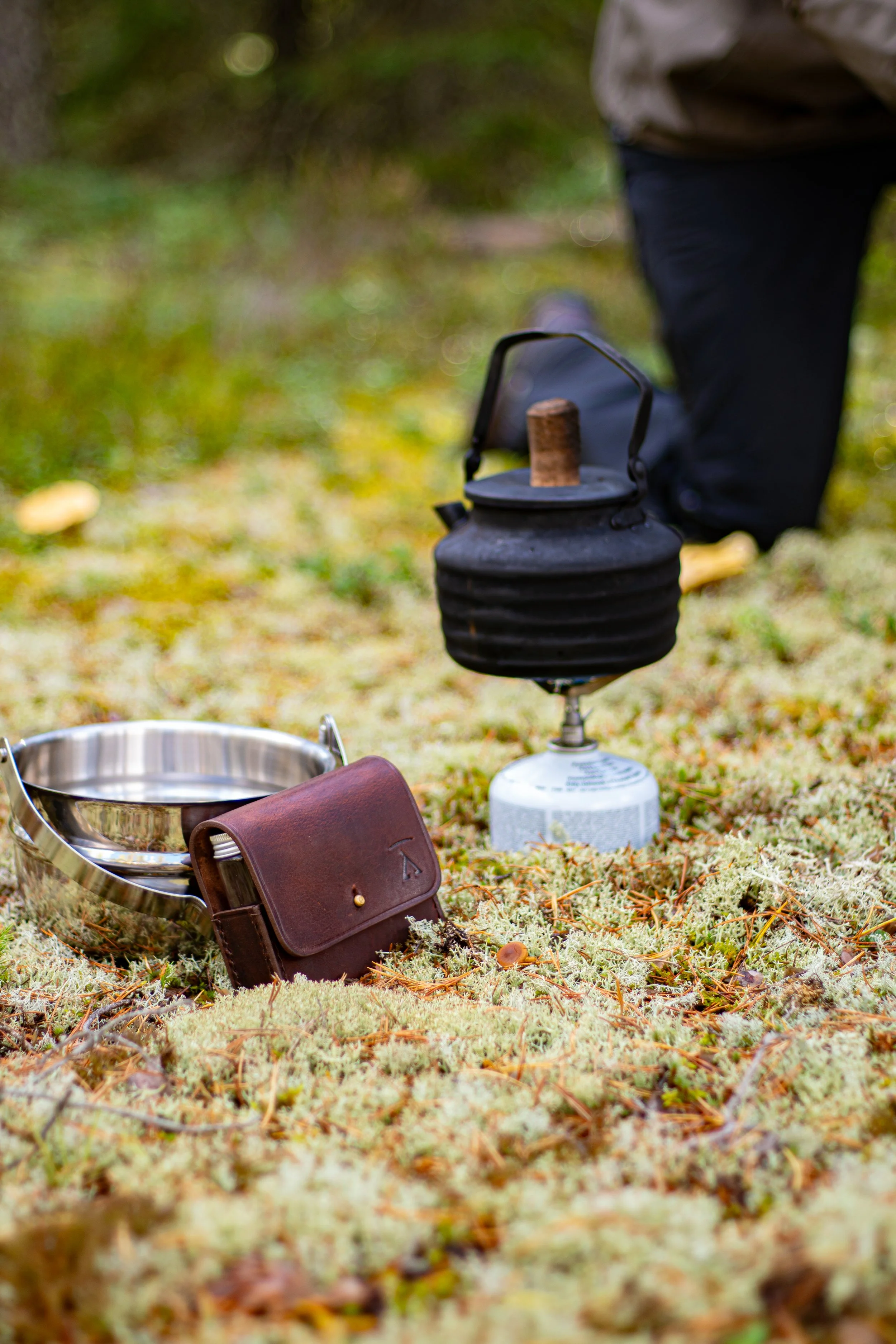 Outdoor camping scene with a vintage black kettle on a portable stove, surrounded by moss, a metal bowl, and a leather pouch, with a blurred background of trees and autumn leaves.