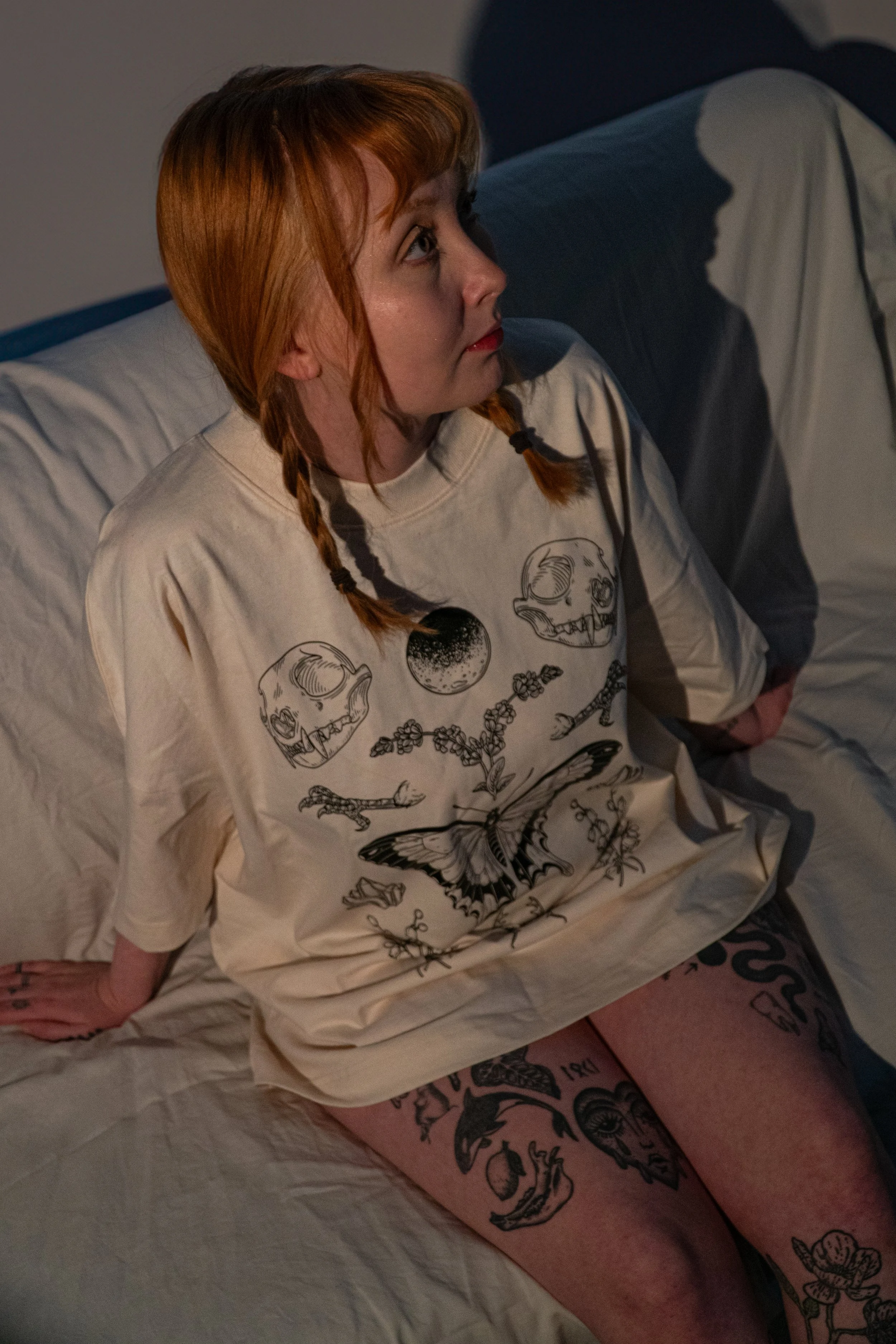 A woman with red hair in braids sitting on a bed, wearing a cream-colored sweatshirt with skull and plant designs, and displaying multiple tattoos on her legs.