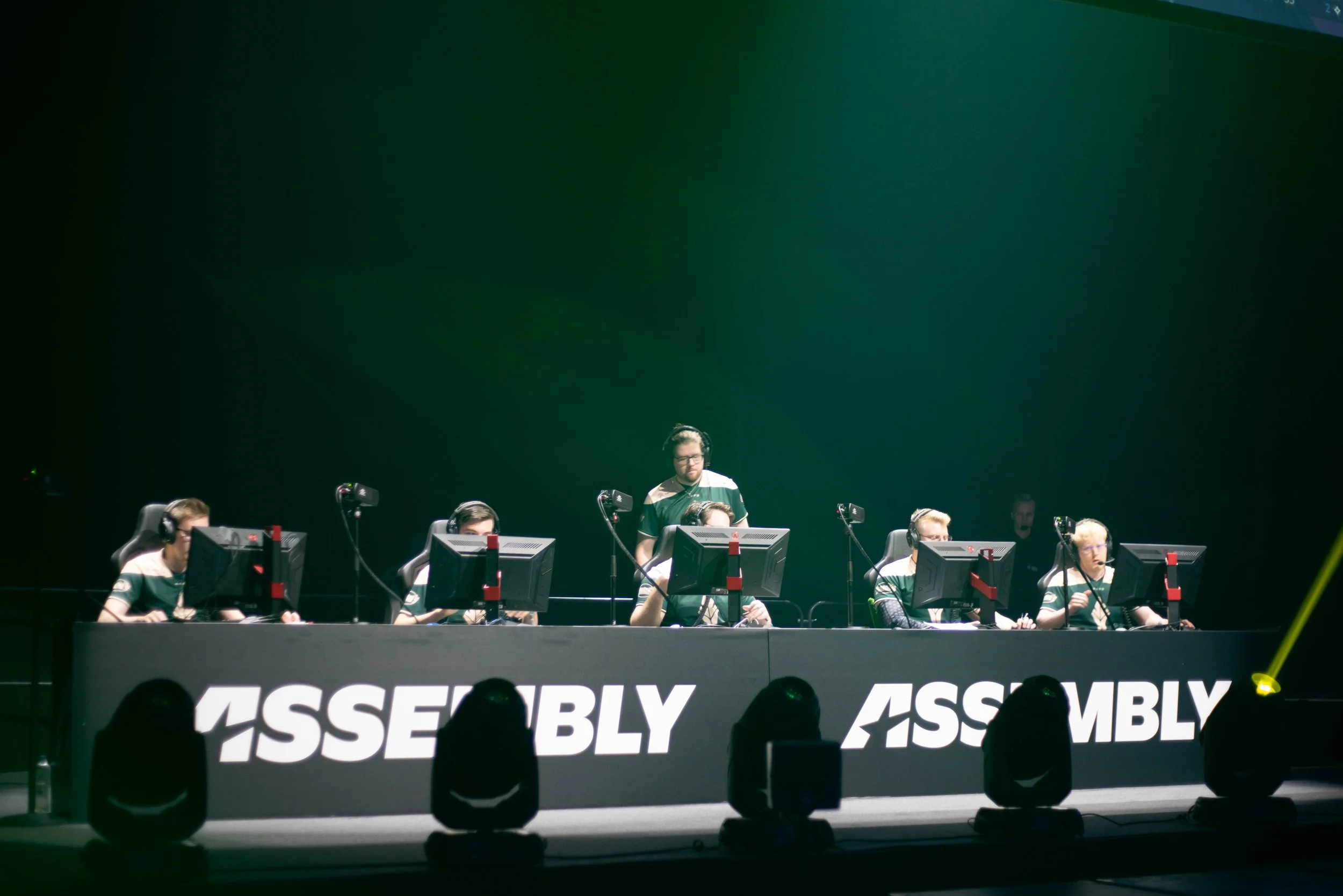 A group of gamers at a professional gaming event, seated at a long table with monitors, wearing headphones, and focused on their screens. The background displays the words 'ASSEMBLY' in large white letters.