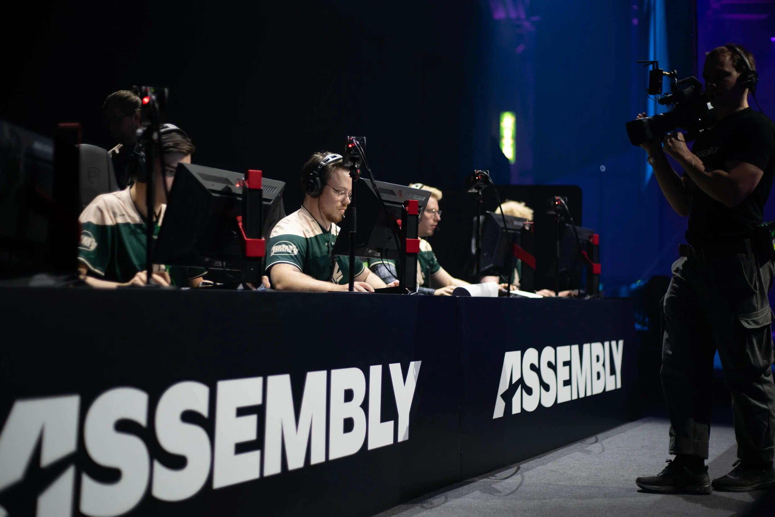 Esports players sitting at computers with headphones, a cameraman filming from the right, and a backdrop with bold white text reading 'ASSEMBLY' in an esports arena.