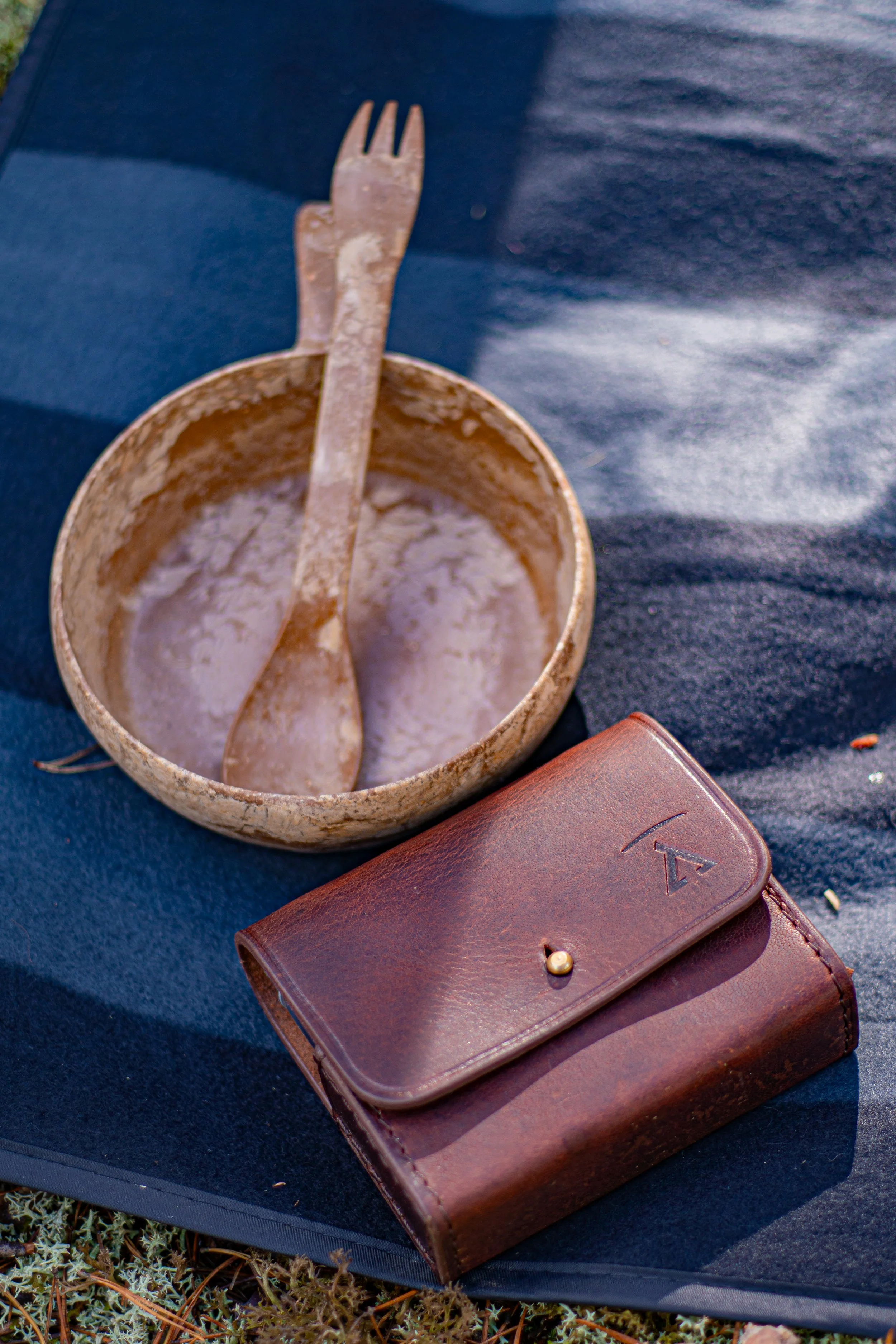 A tin cup with remnants of a drink and a metal fork inside sits on a dark surface. Beside it is a closed brown leather wallet with a gold snap button and an embossed emblem, also on the dark surface.
