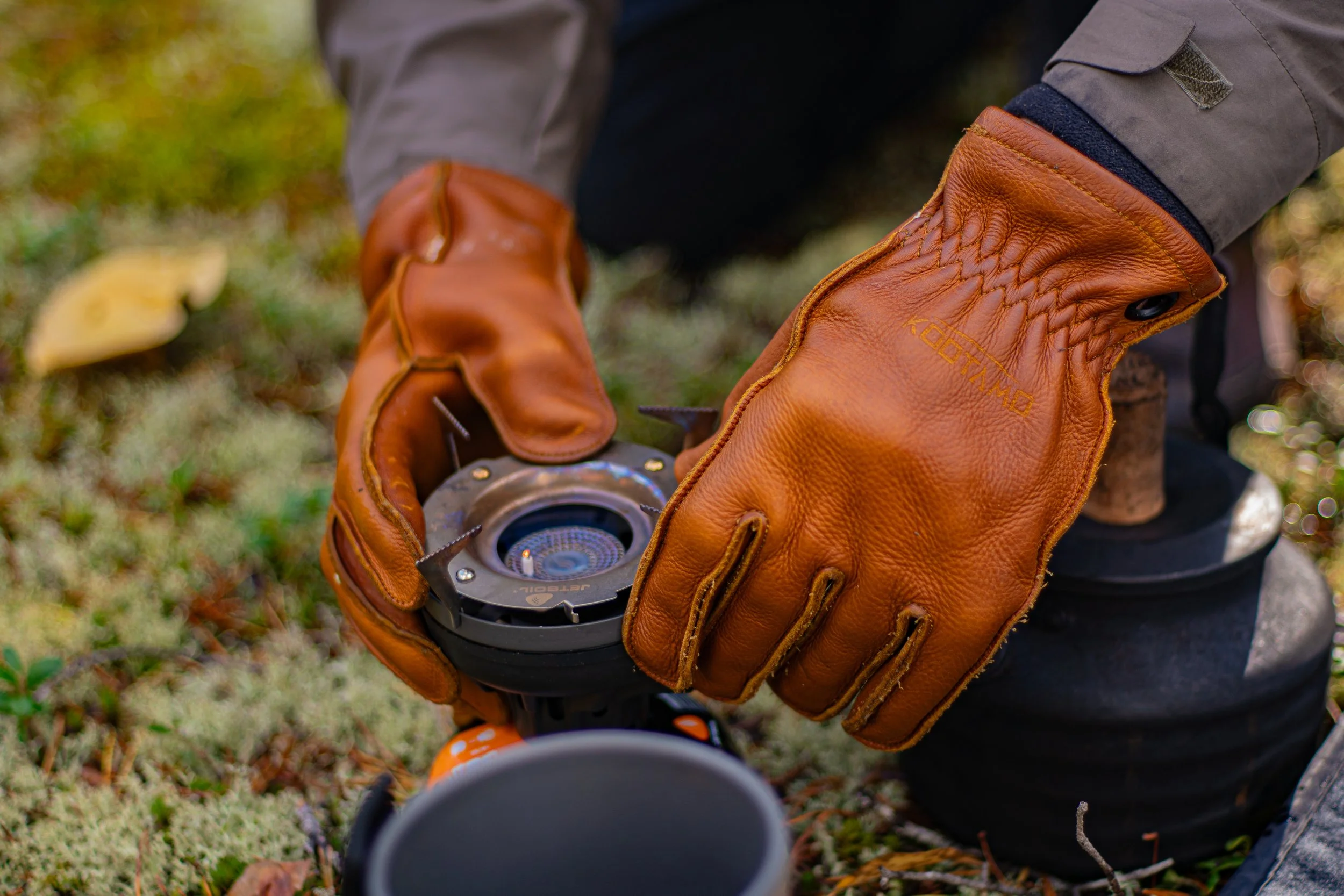 Person wearing brown leather gloves holding a camera lens or a flashlight outdoors on grass with fallen leaves.