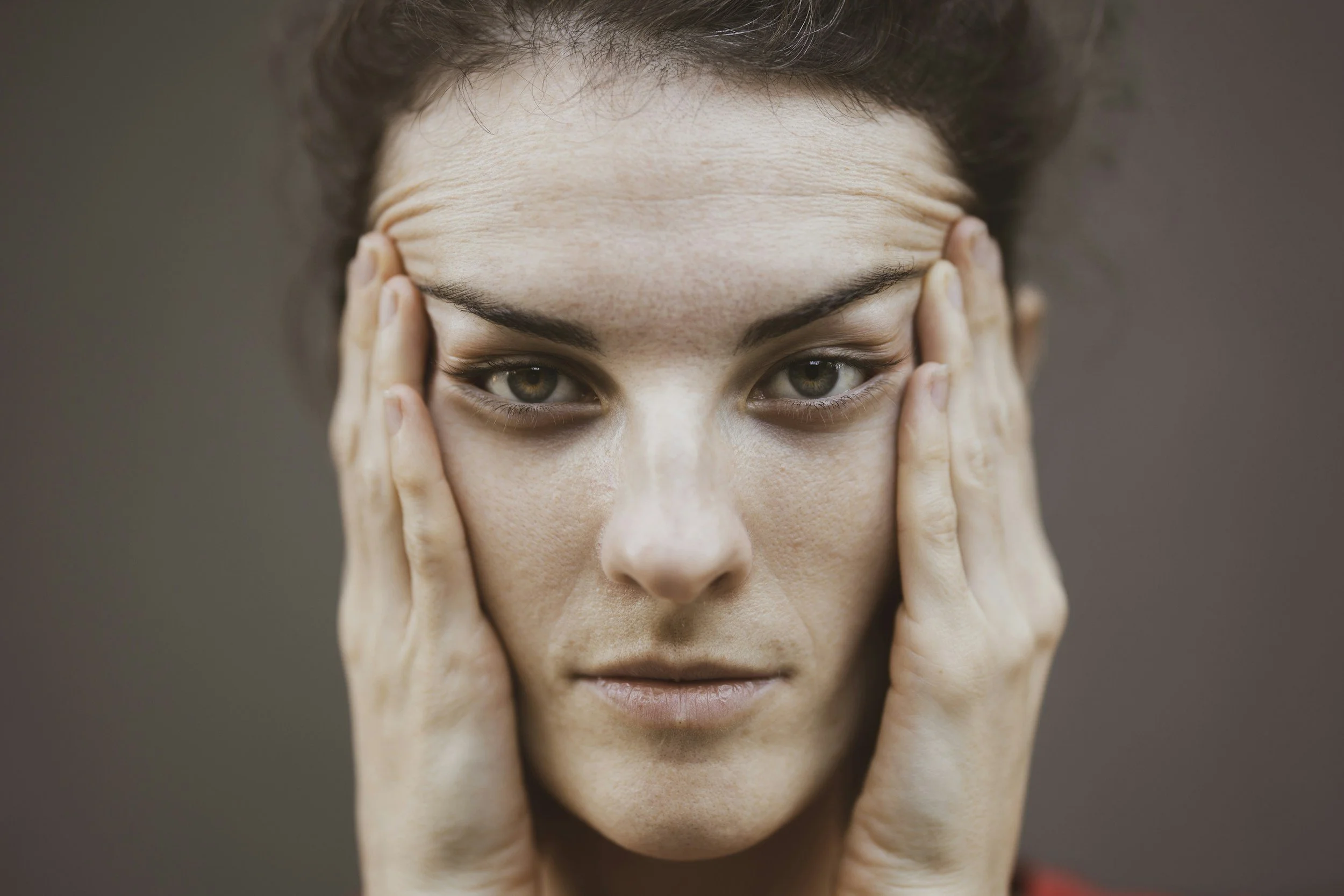 A young woman with her face held gently by her hands, with her fingers touching her temples, looking directly at the camera with a calm expression.