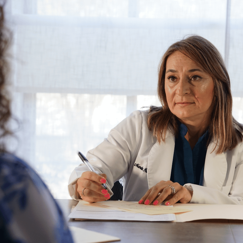 A woman in a white coat speaking with a patient at a desk, holding a pen and taking notes, in a well-lit room with large windows.