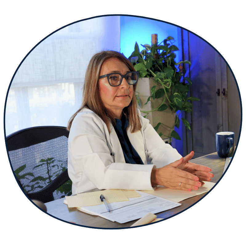 A woman with shoulder-length brown hair, wearing glasses and a white blazer, sitting at a desk with papers, a pen, a mug, and a large green plant in the background.