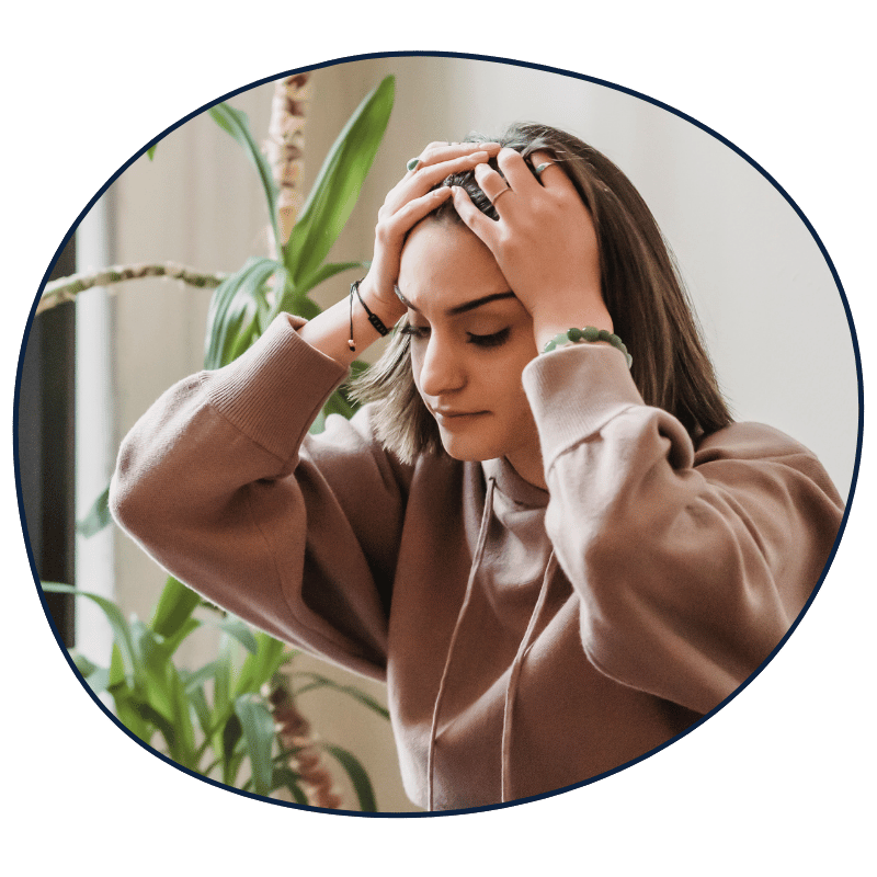 A young woman experiencing stress or headache, holding her head with both hands, sitting indoors near a plant.