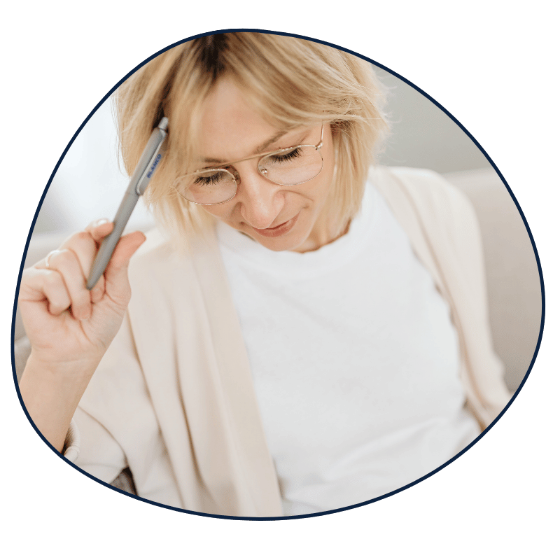 A woman with blonde hair and glasses appears to be deep in thought, holding a pen to her temple.