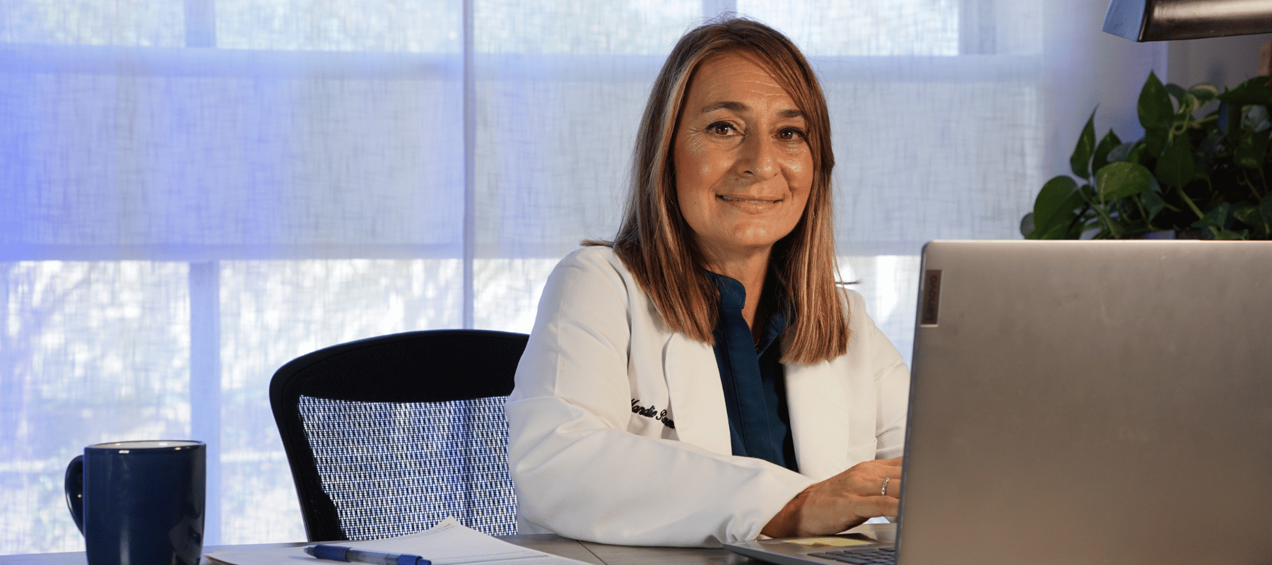 A woman with light brown hair wearing a white lab coat sitting at a desk with a laptop, a pen, and a notebook, smiling at the camera in a well-lit office with sheer curtains and a potted plant.