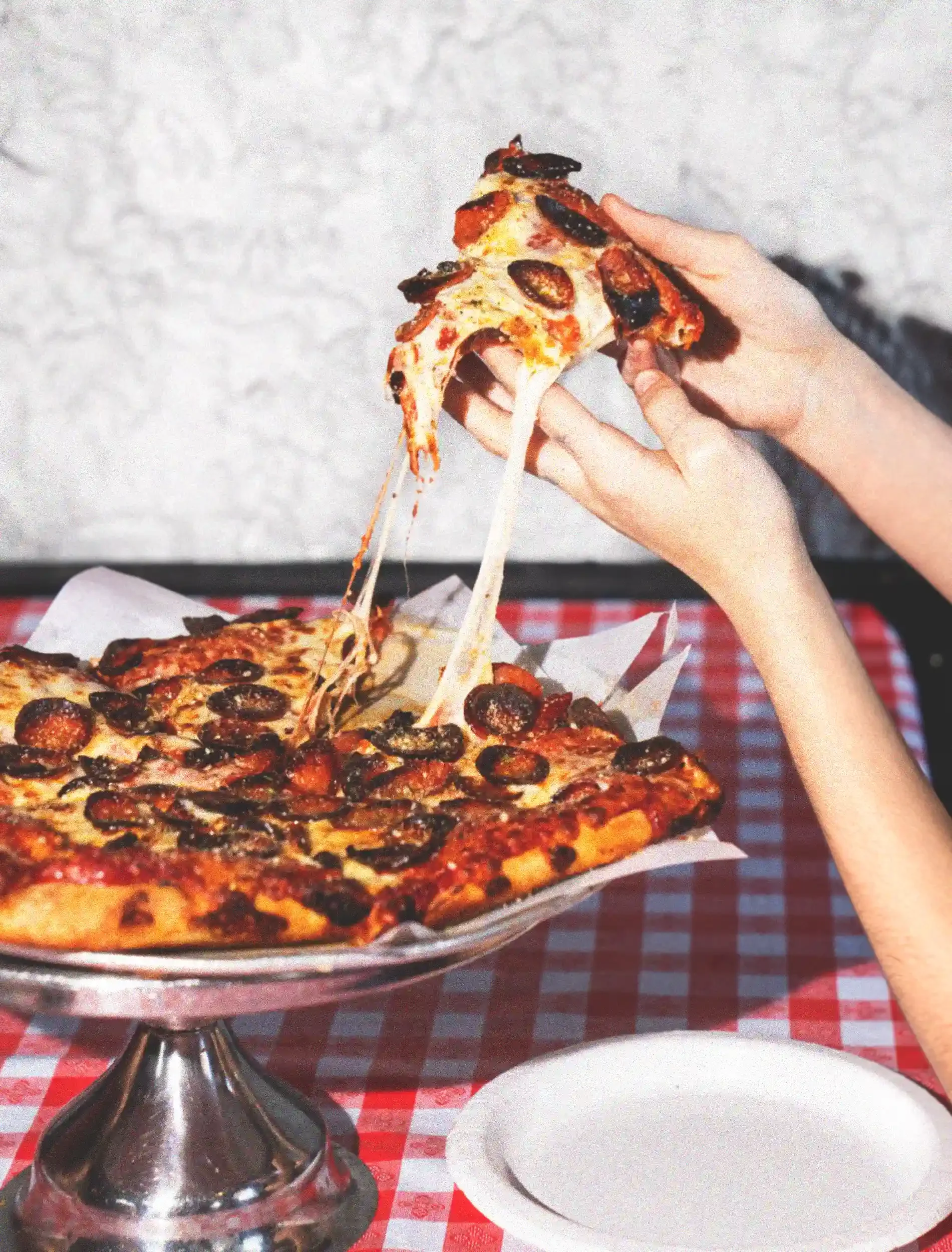 A person holding a slice of pepperoni and black olive pizza with melted cheese dripping. There is a whole pizza on the table with the same toppings, and a white plate on a red-checked tablecloth.