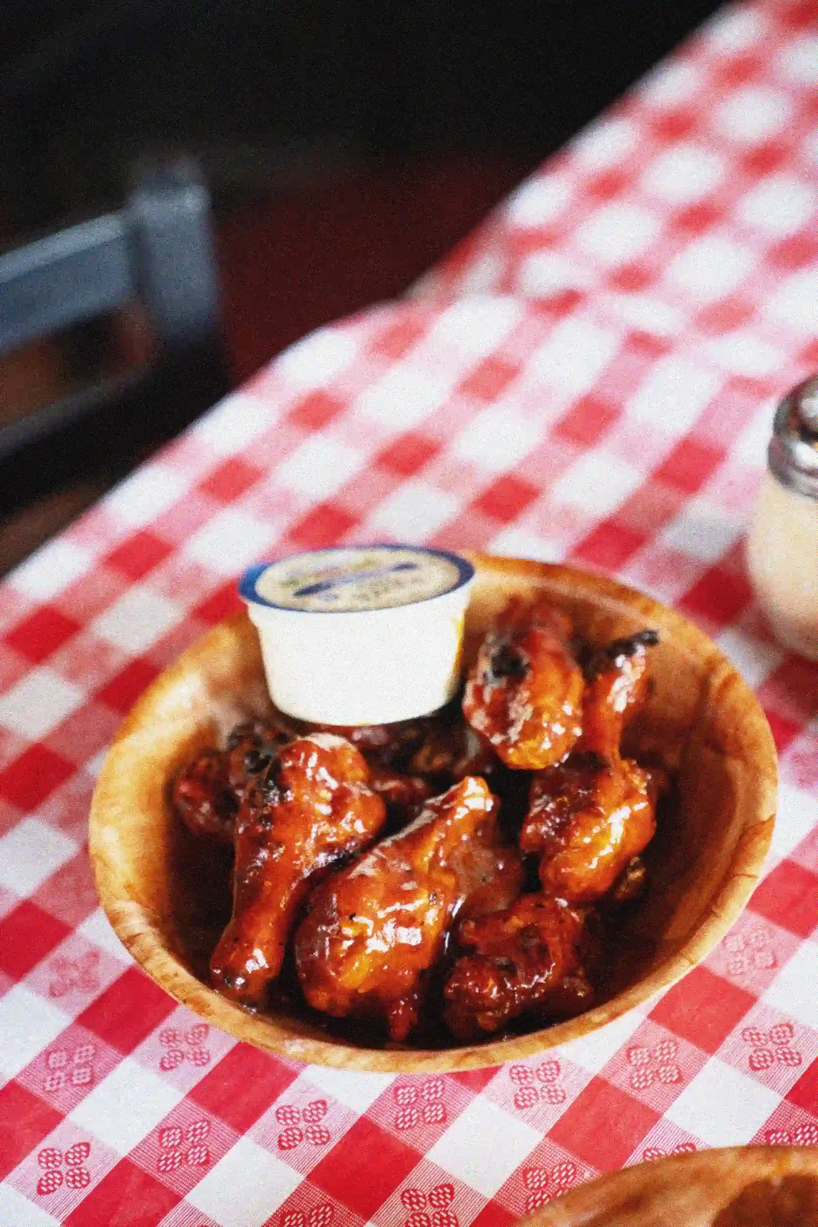 A bowl of boneless chicken wings covered in barbecue sauce placed on a red and white checkered paper tablecloth.