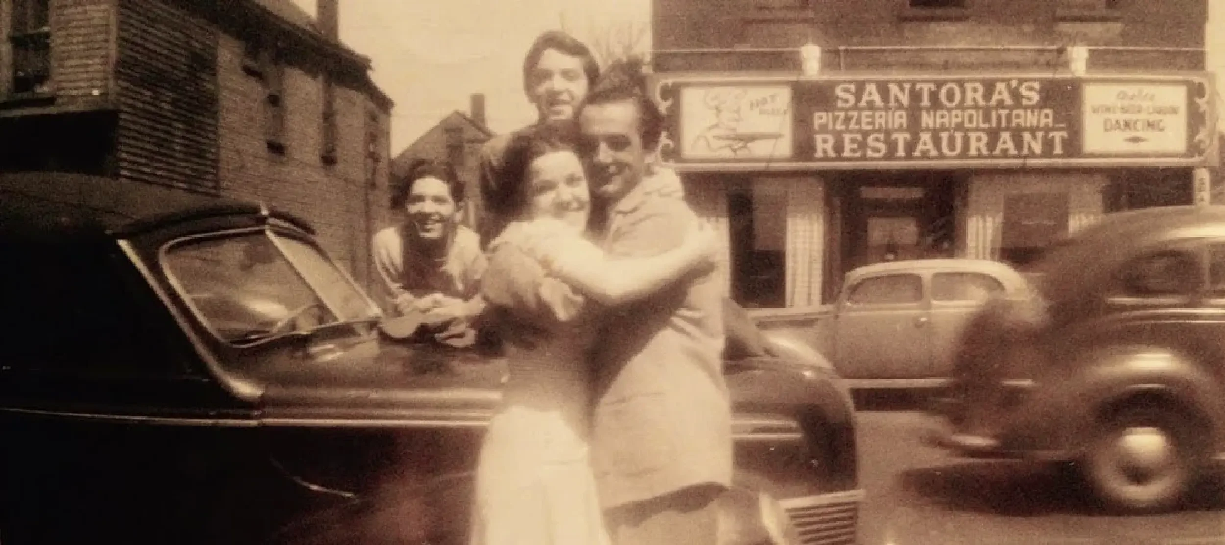 Vintage black-and-white photo of four smiling people posing on a city street in front of a restaurant sign that reads 'Santora's Pizzeria Napolitana Restaurant.' Two cars are visible in motion.