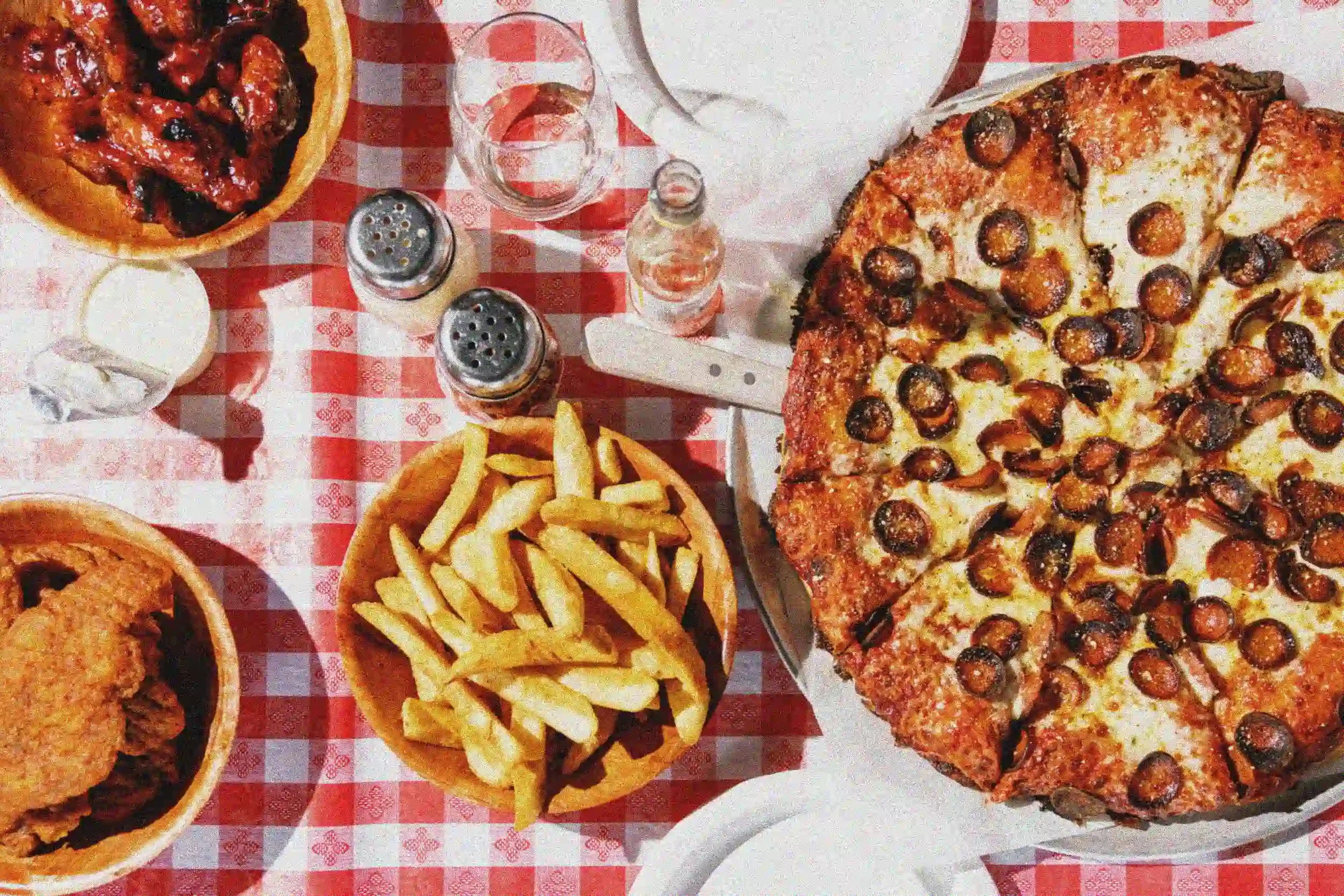 A table with a variety of foods including two large pepperoni pizzas, French fries, a plate of chicken wings, a bowl of barbecue ribs, salt and pepper shakers, and bottles of condiments on a red checkered tablecloth.