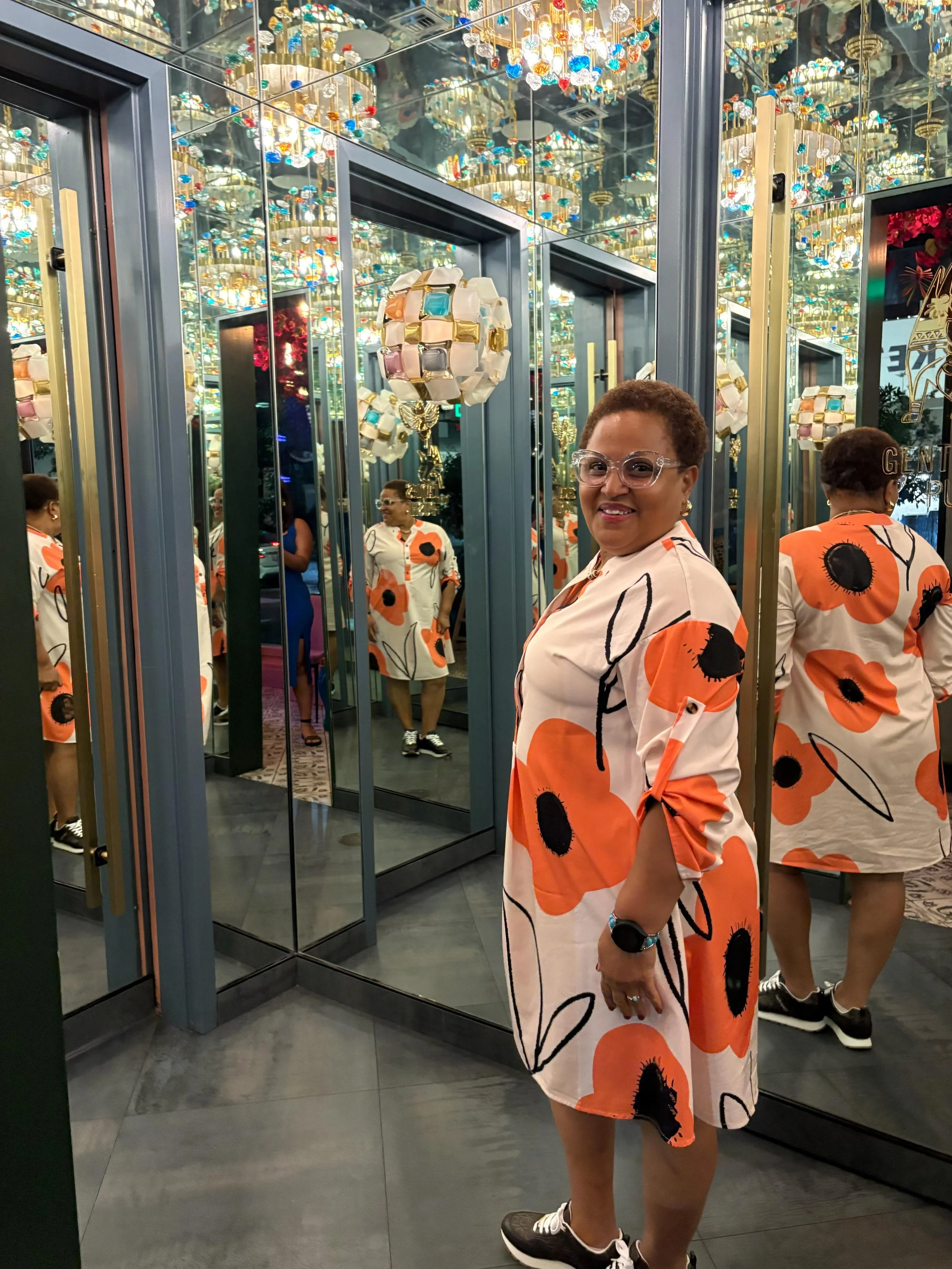 Mujer de cabello rizado, gafas transparentes, vestido con estampado de flores grandes negras y naranjas, en una tienda con espejos y lámparas decorativas, sonriendo hacia la cámara.