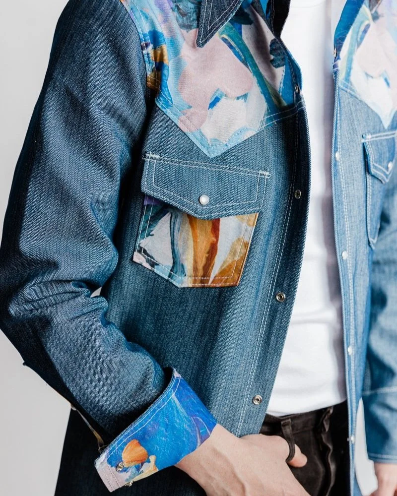 Close-up of a denim jacket with colorful abstract patterns on the collar, chest pockets, and cuffs, worn over a white shirt.